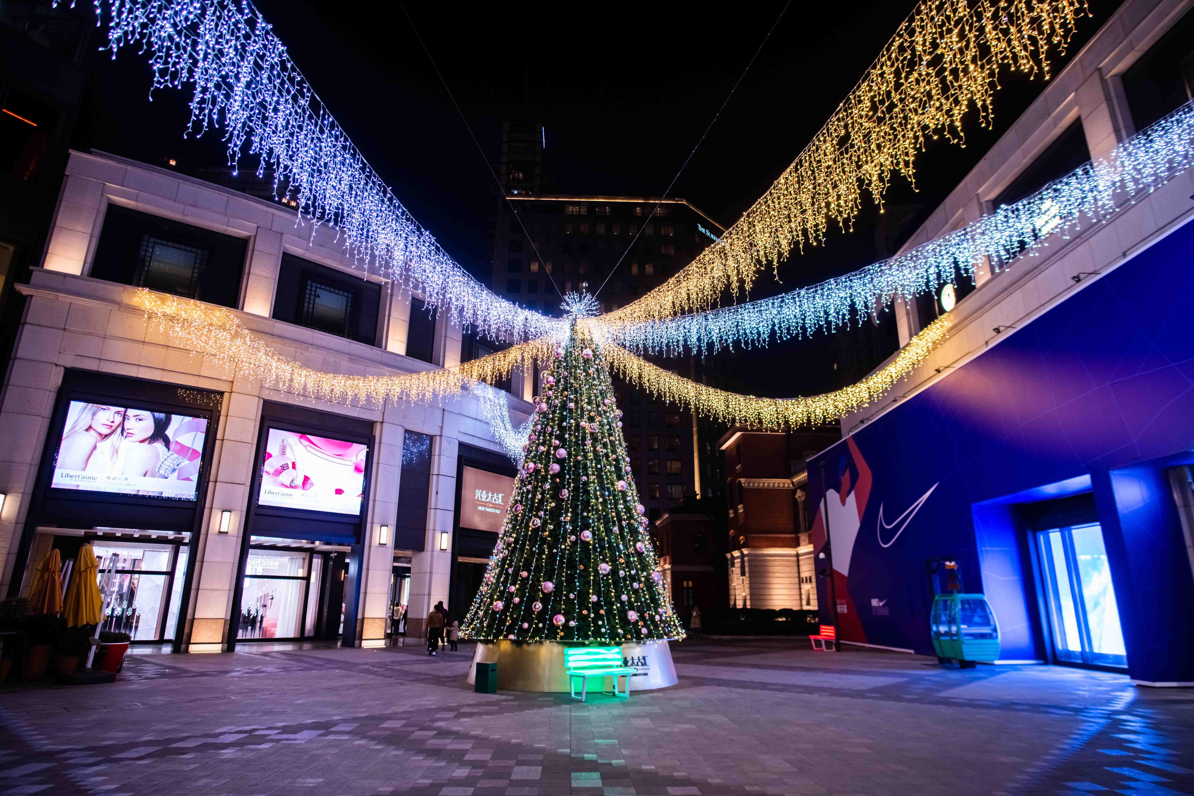 Weihnachtlich beleuchteter Platz mit großem Tannenbaum in der Mitte, umgeben von Geschäften und hängenden Lichterketten.