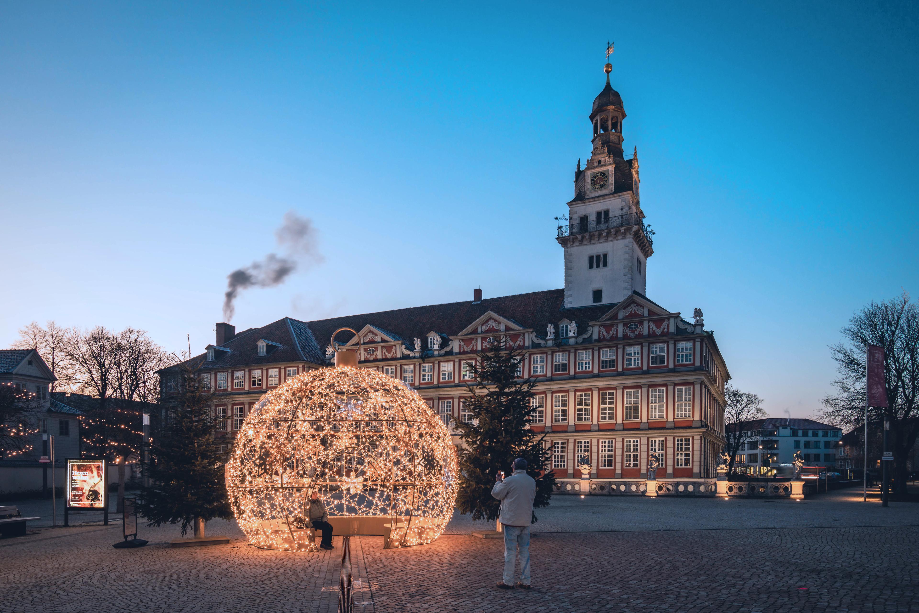 Große leuchtende Kugelinstallation auf einem Platz vor einem historischen Gebäude in der Dämmerung, gesehen von zwei Personen.