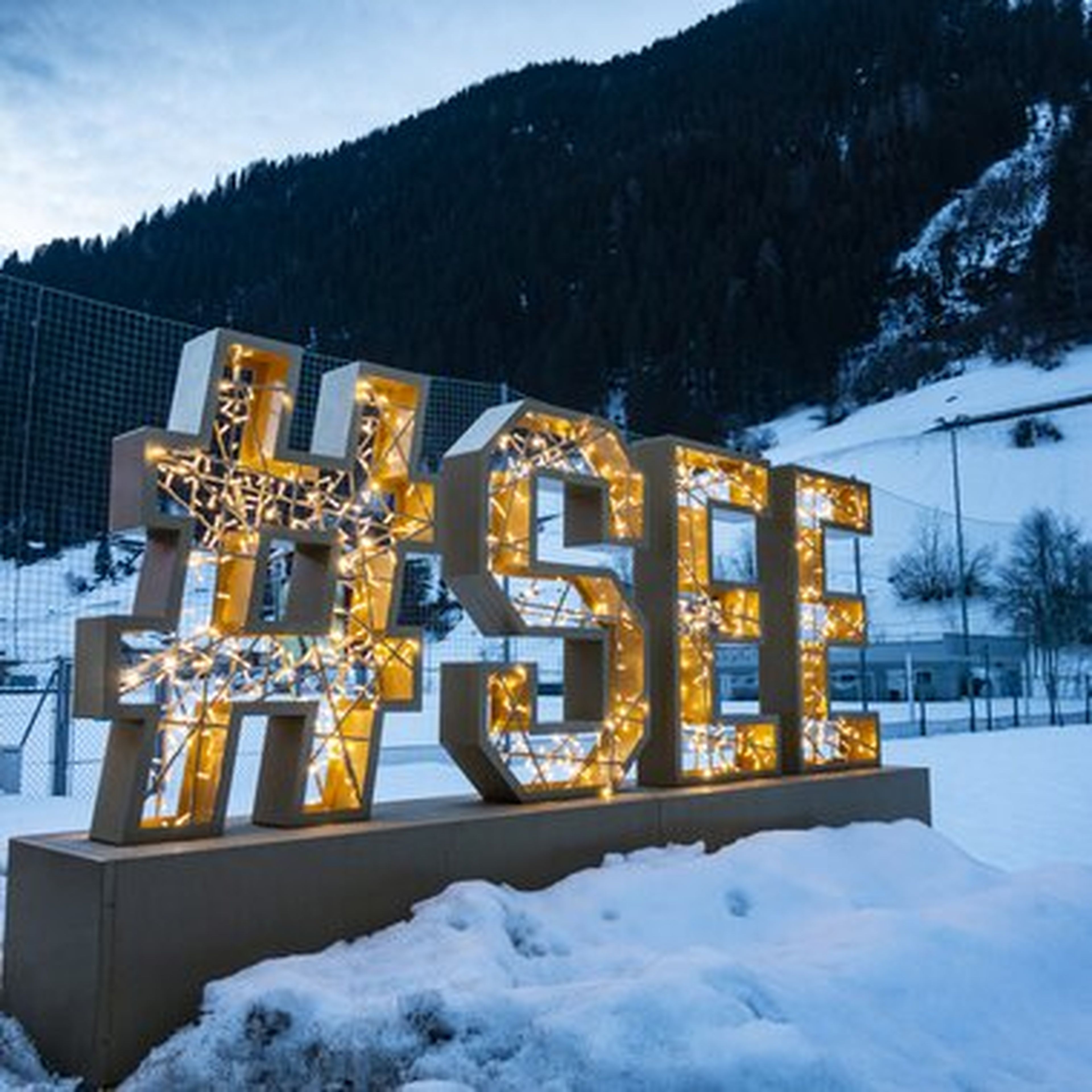 Großes, golden leuchtendes #SEE-Schild steht im winterlichen Schnee vor einer verschneiten Bergkulisse unter dunklem Himmel.