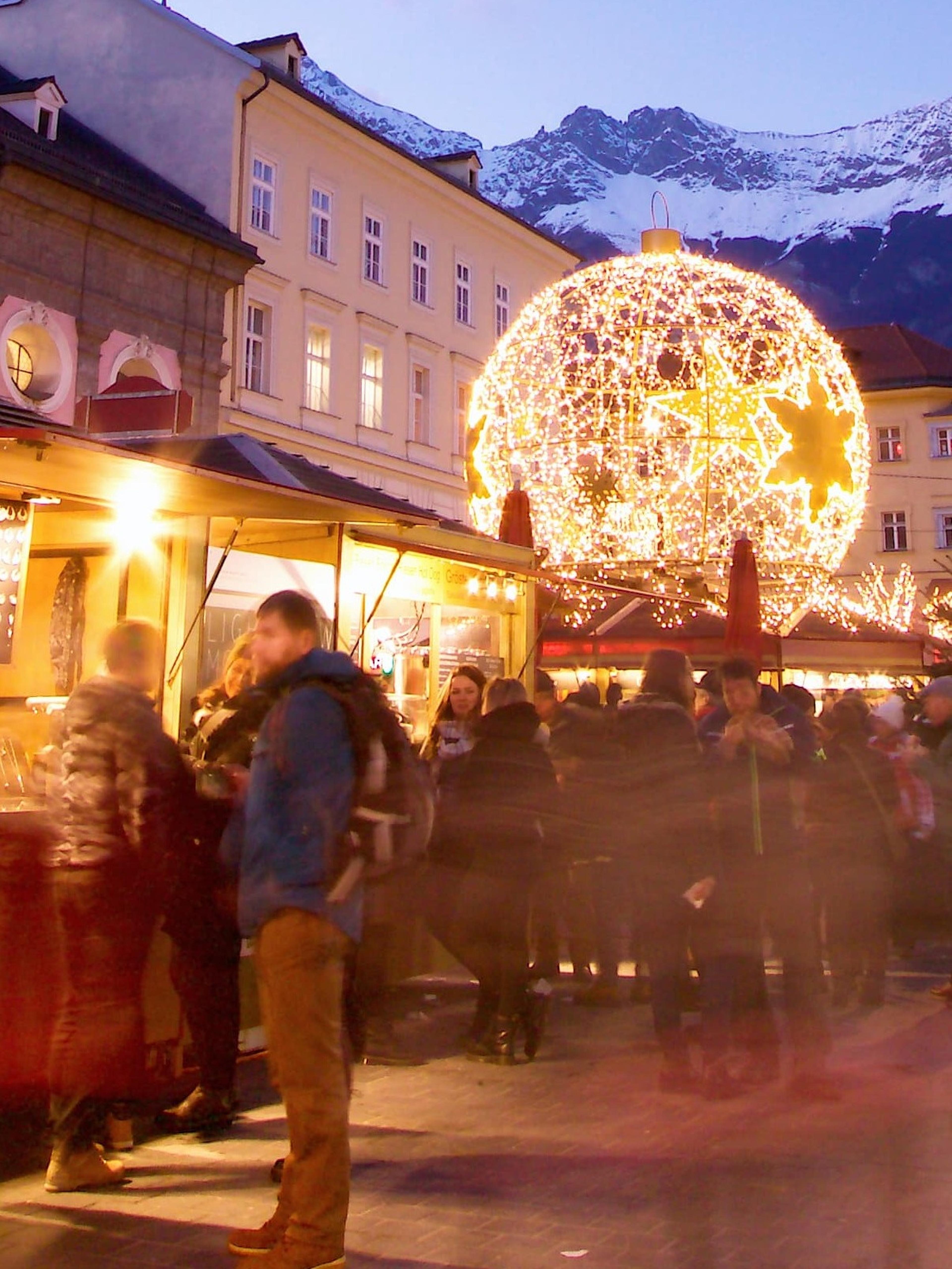 Festive street market at dusk in a European town with a clock tower, lit globe decoration, shops, and blurred crowd, with snowy mountains in the background.