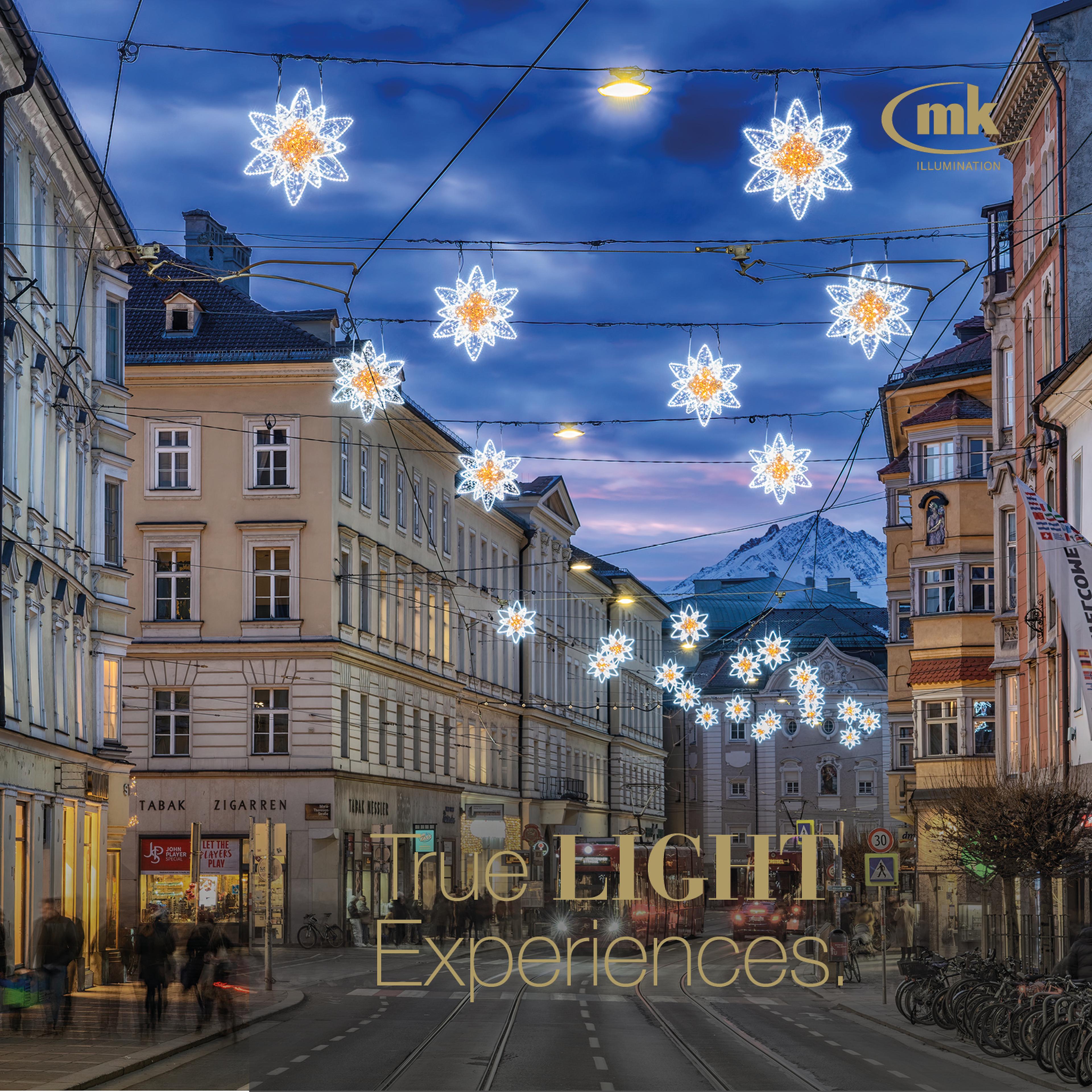Beleuchtete Sternendekoration über einer städtischen Straße bei Abenddämmerung, umgeben von historischen Gebäuden und Bergen im Hintergrund.
