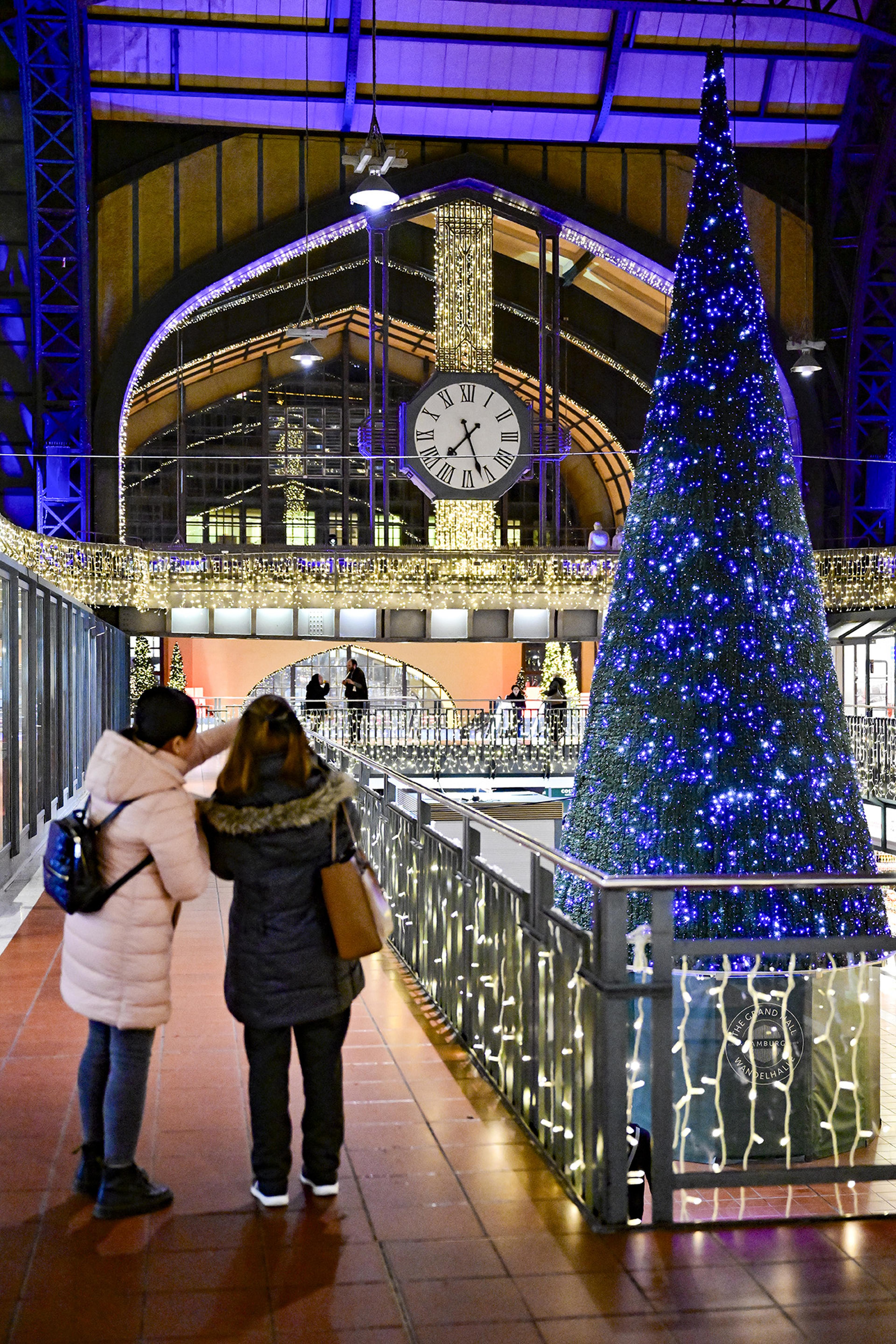 Personen stehen vor einem großen, blau beleuchteten Weihnachtsbaum in einem festlich dekorierten Bahnhof mit einer großen Uhr.