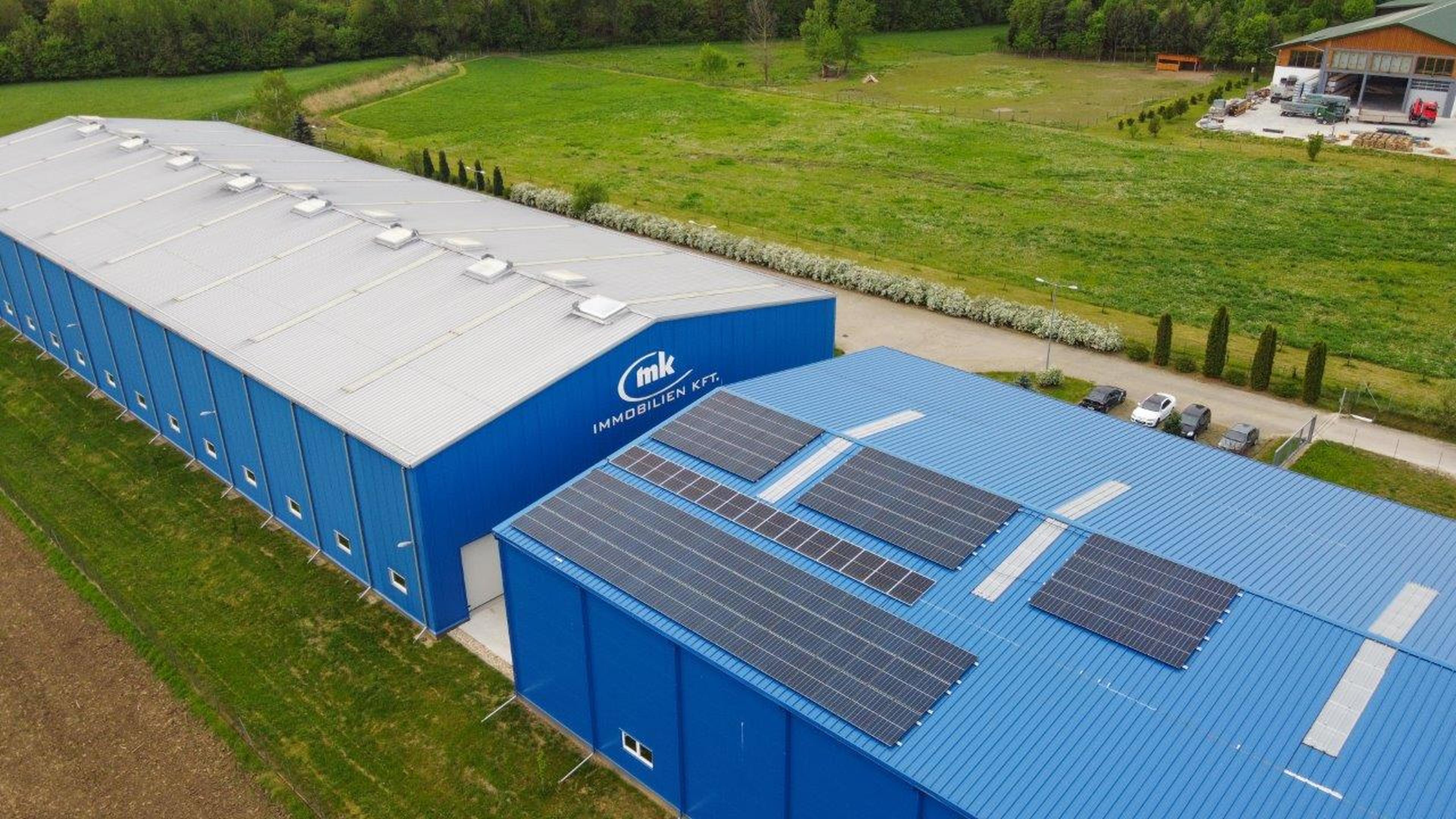 Ein großes blaues Gebäude mit Solarpanelen auf dem Dach, umgeben von grünen Feldern und Bäumen.