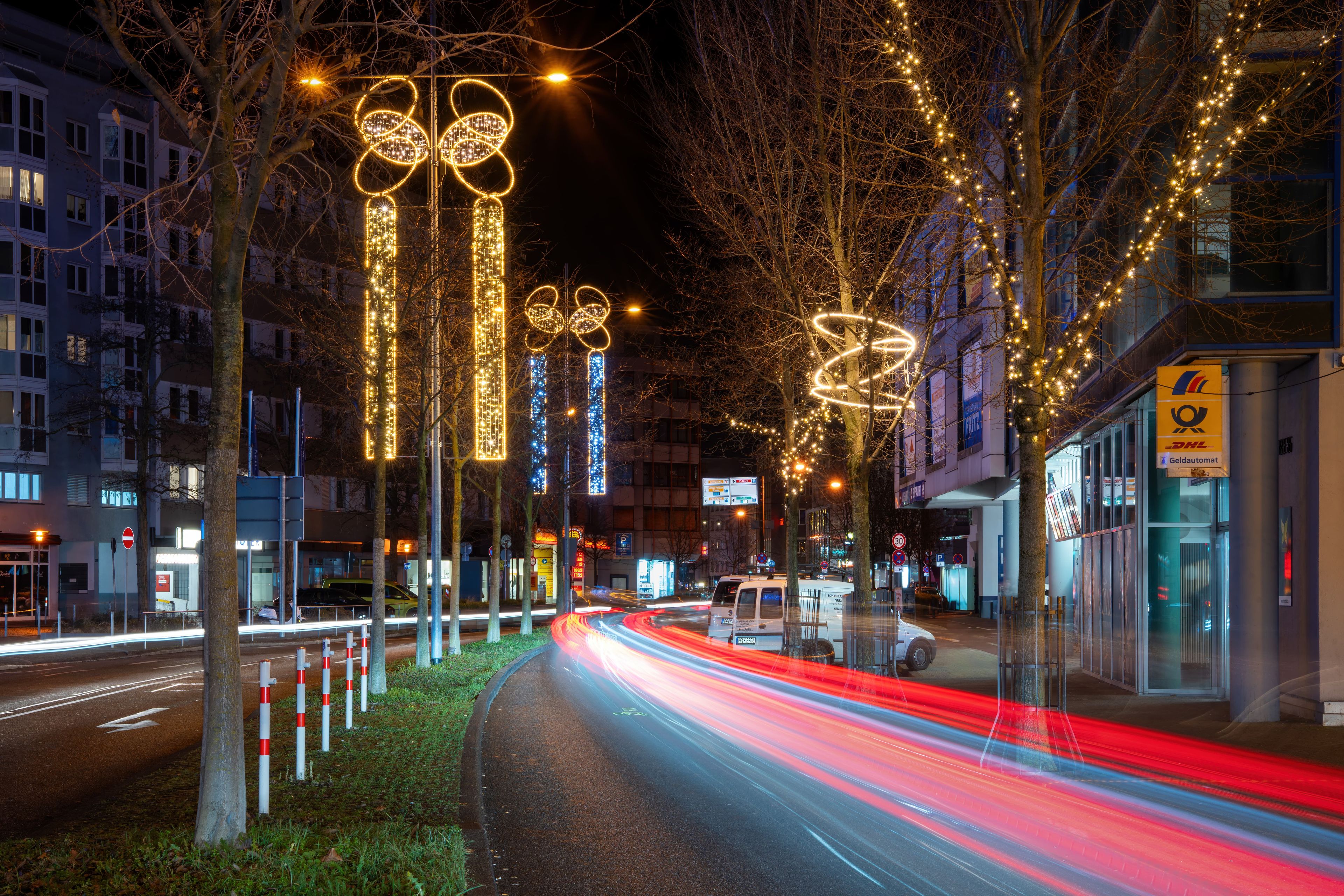 City street at night with festive lights in tree shapes, glowing decorations, and long-exposure car light trails on a curving road.