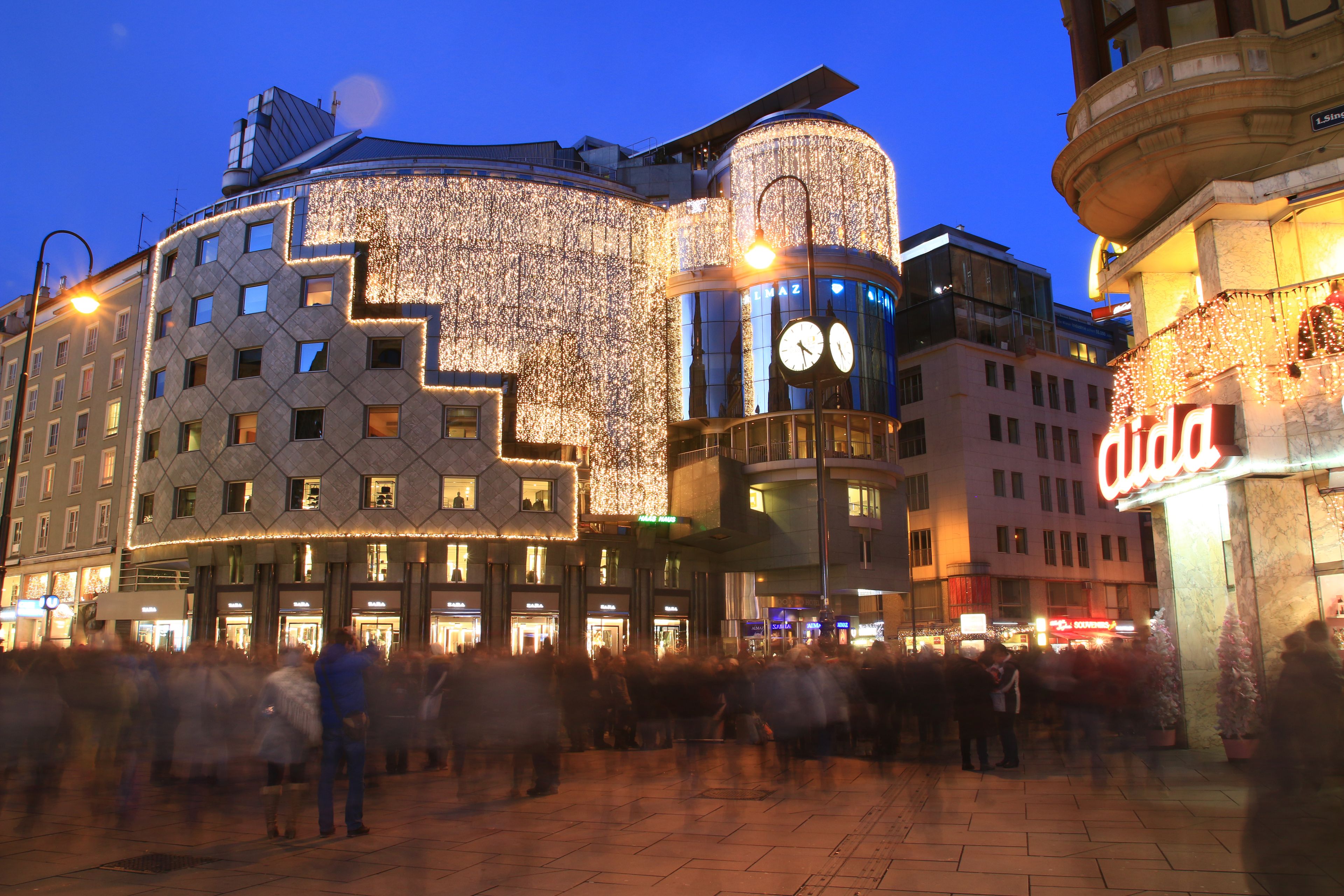 Belebter Abend in der Stadt, modern beleuchtete Architektur mit festlichen Lichterketten und Schaufenstern, Menschen flanieren im Vordergrund.
