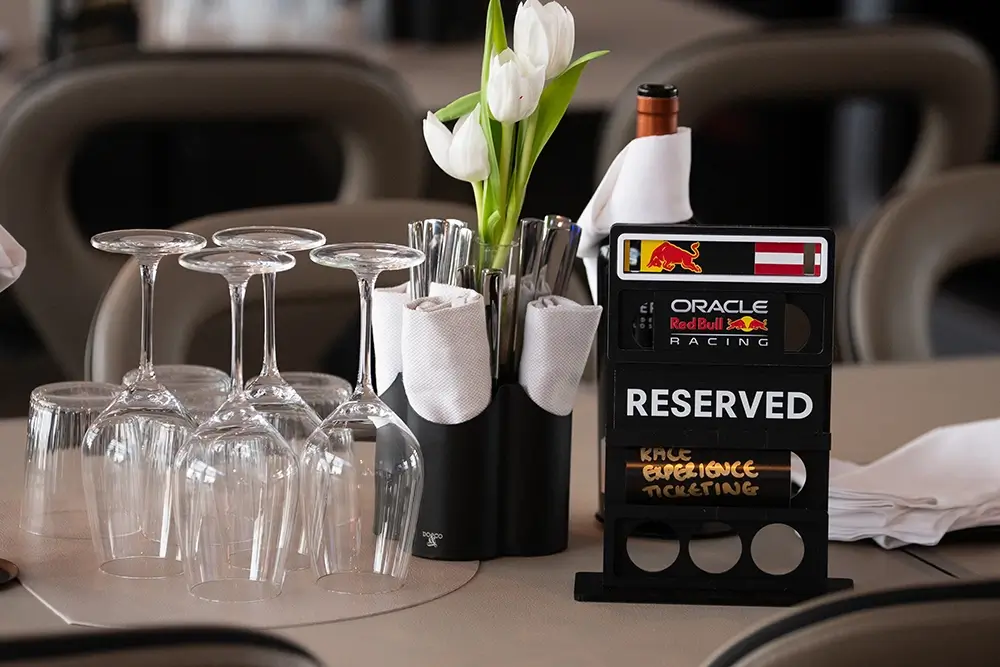 Table setting with four upside-down wine glasses, white tulips, cutlery in napkins, and a "reserved" sign featuring racing brand logos.