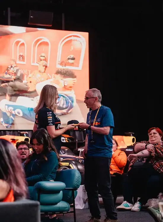 People watching a F1 race on a large screen in MK-7 event space. A woman and man stand talking in the foreground.