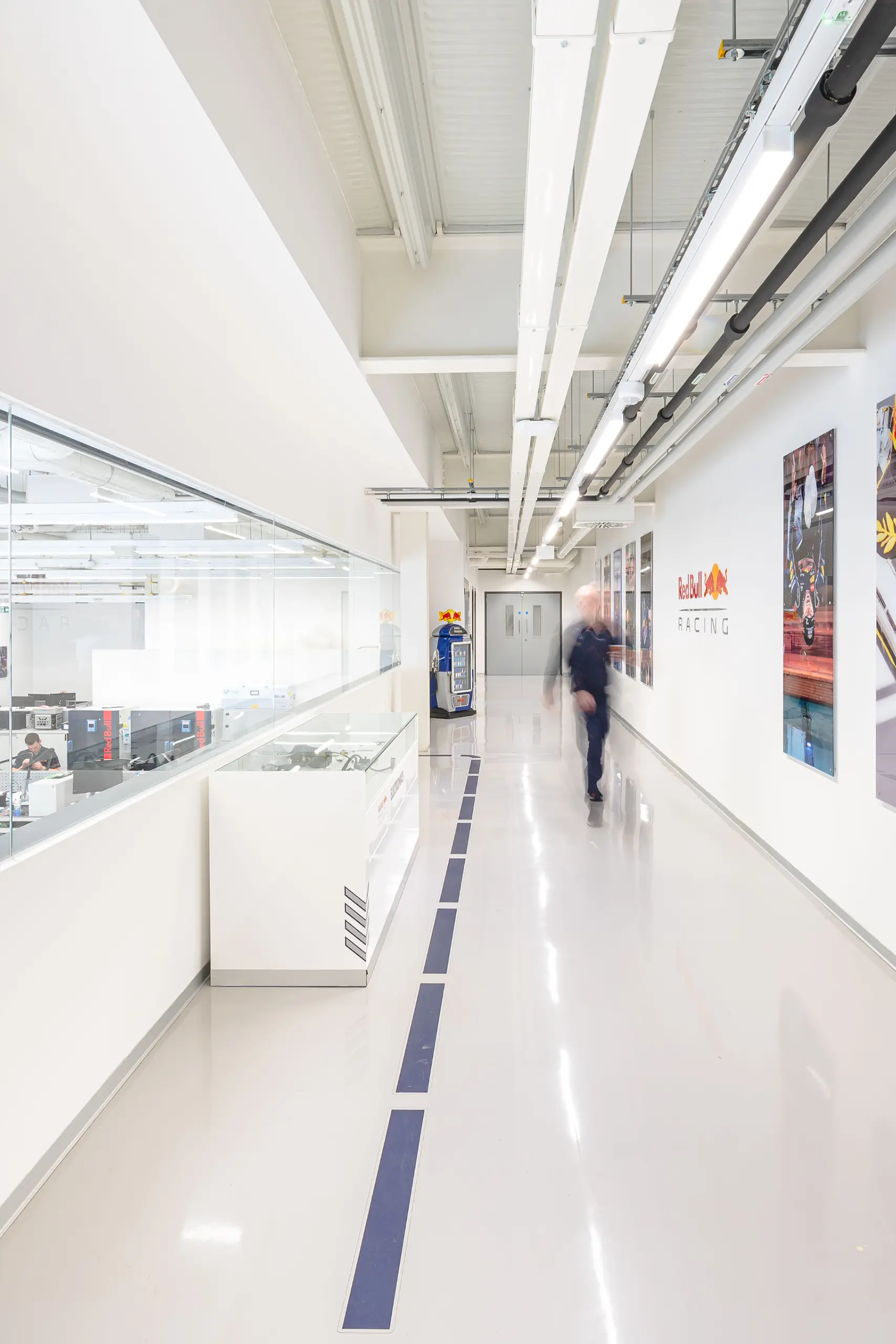 Bright, modern hallway with glass wall displaying workstations and Red Bull Racing posters. A blurred person walks along the polished floor.
