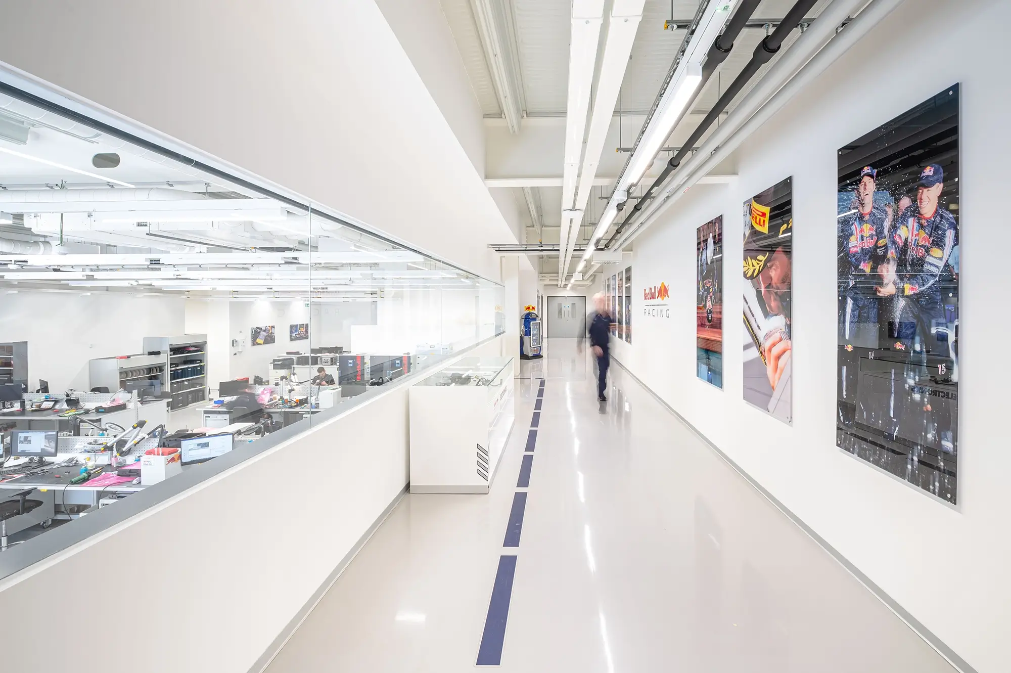 Bright office corridor with glass walls showing workstations, racing team posters on the walls, and a person walking by.