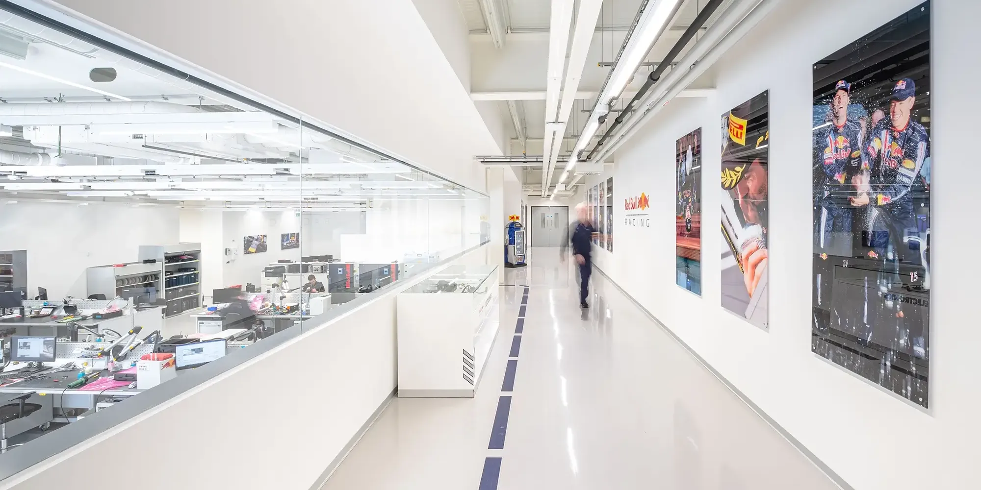 Bright office corridor with glass walls showing workstations, racing team posters on the walls, and a person walking by.