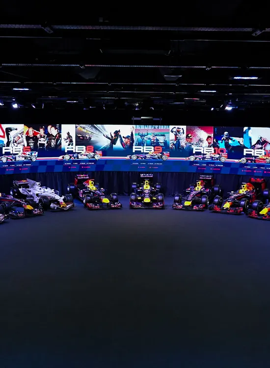 A collection of Red Bull Racing Formula 1 cars displayed in a semicircle, with large screens showing racing imagery above them.