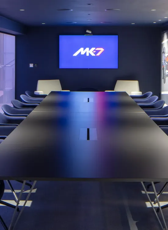 Modern conference room with a long table, blue chairs, mounted screen displaying "MK7," and shelves with racing helmets.