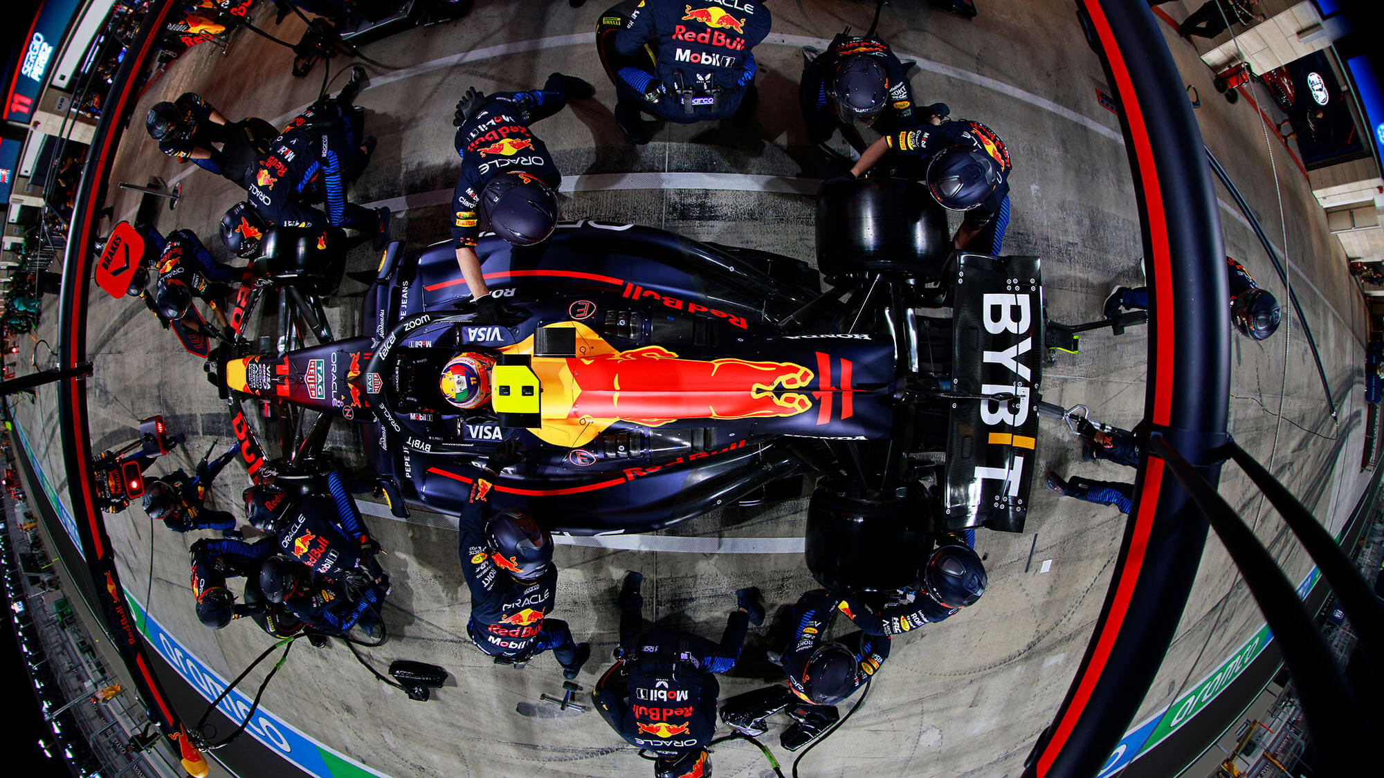 Aerial view of a Formula 1 pit stop with team members surrounding a race car for service, brightly lit under night sky conditions.