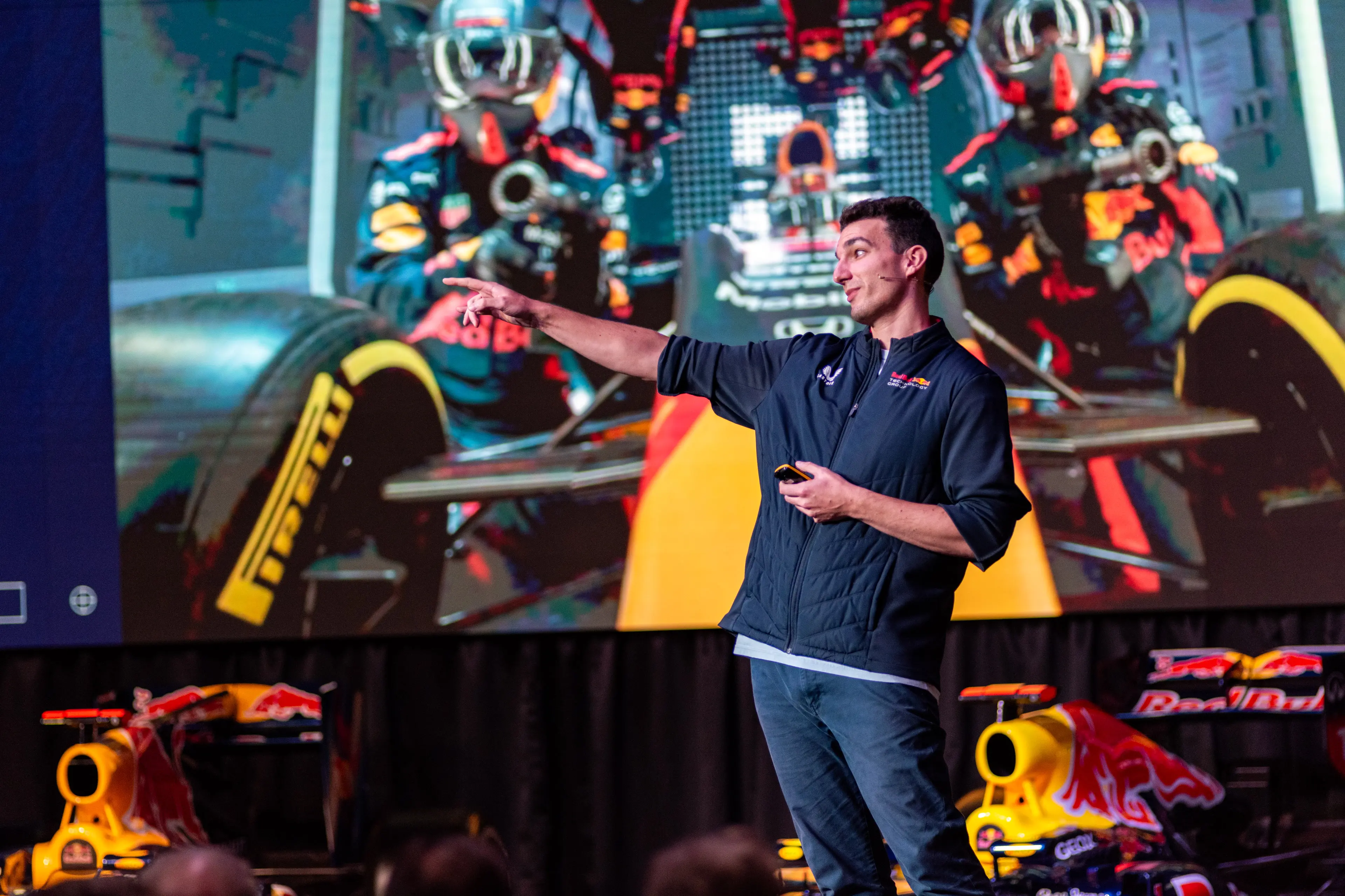 Man presenting on stage with Red Bull racing cars and pit crew image in the background.