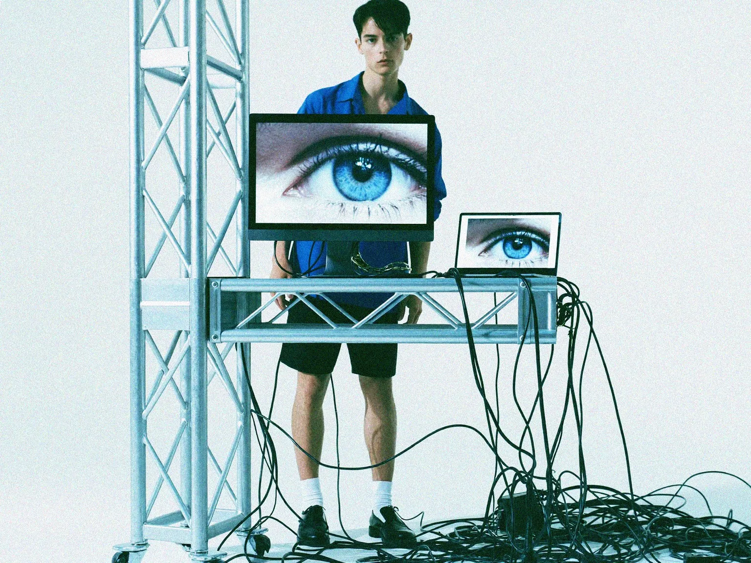 A person behind a screen displaying eyes, surrounded by cables and a metal frame.
