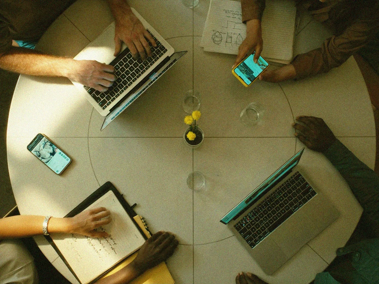 Overhead view of a shared worktable with laptops, notes and several hands.