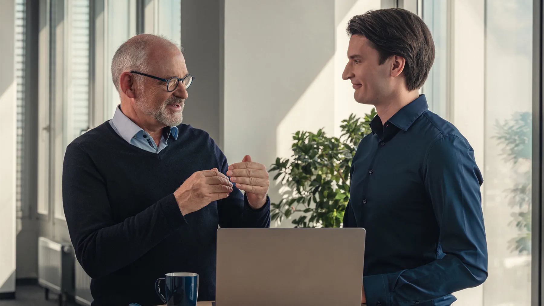 Zwei Männer unterhalten sich in einem modernen Büro vor einem Laptop stehend. Sie lächeln und wirken freundlich.