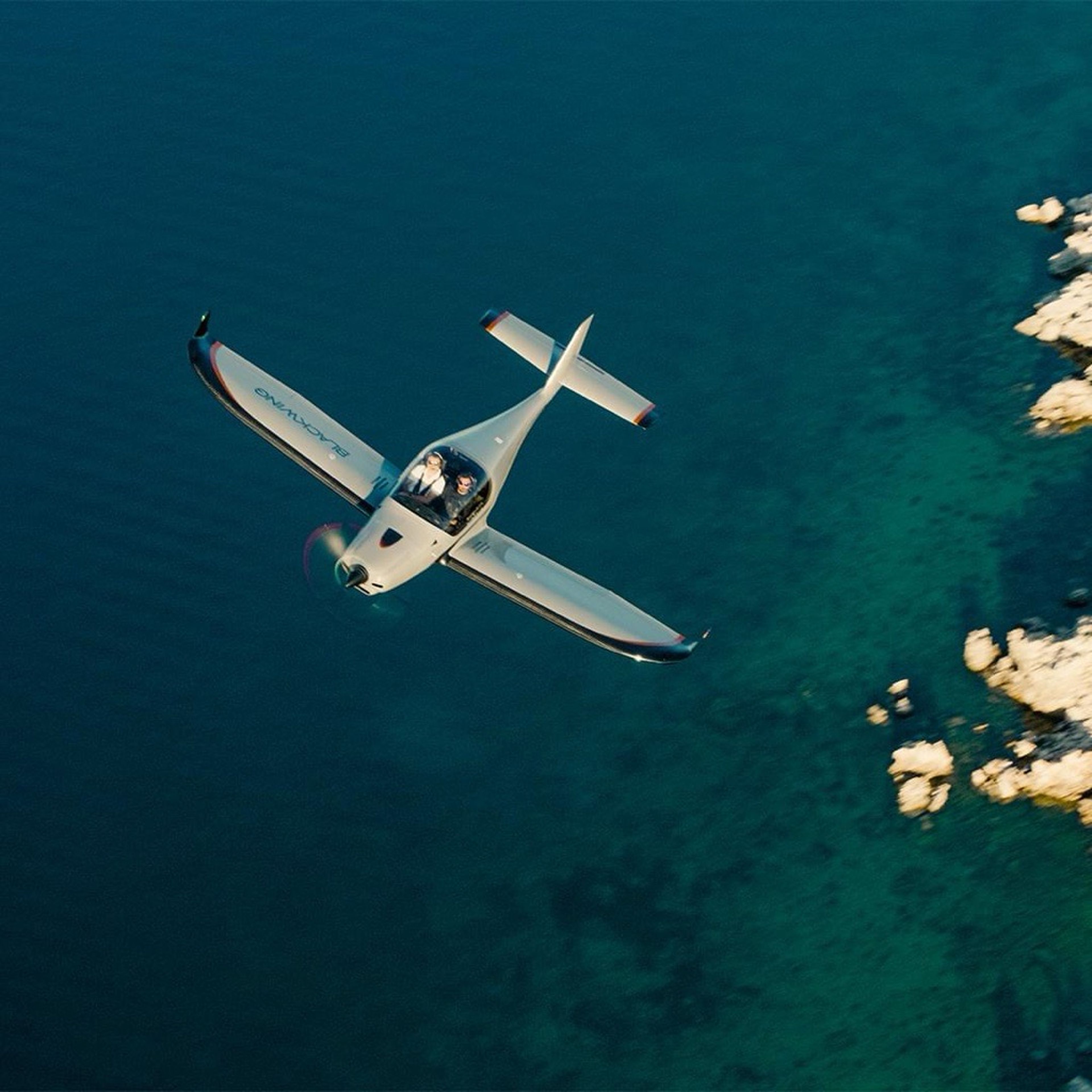 Small airplane flying over clear blue water with rocky shoreline visible to the right.