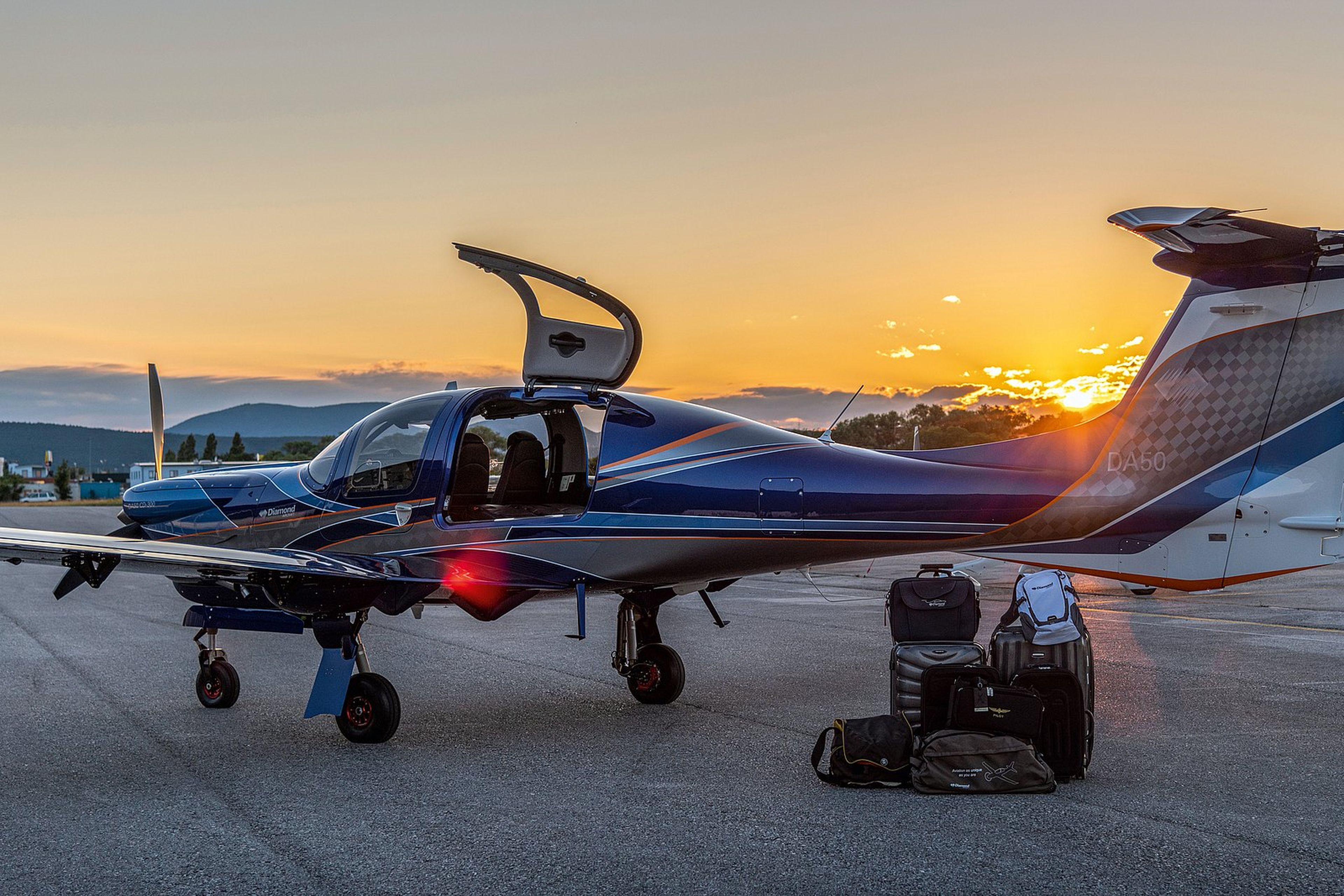 A small blue aircraft with an open door sits on a runway at sunset, surrounded by luggage.