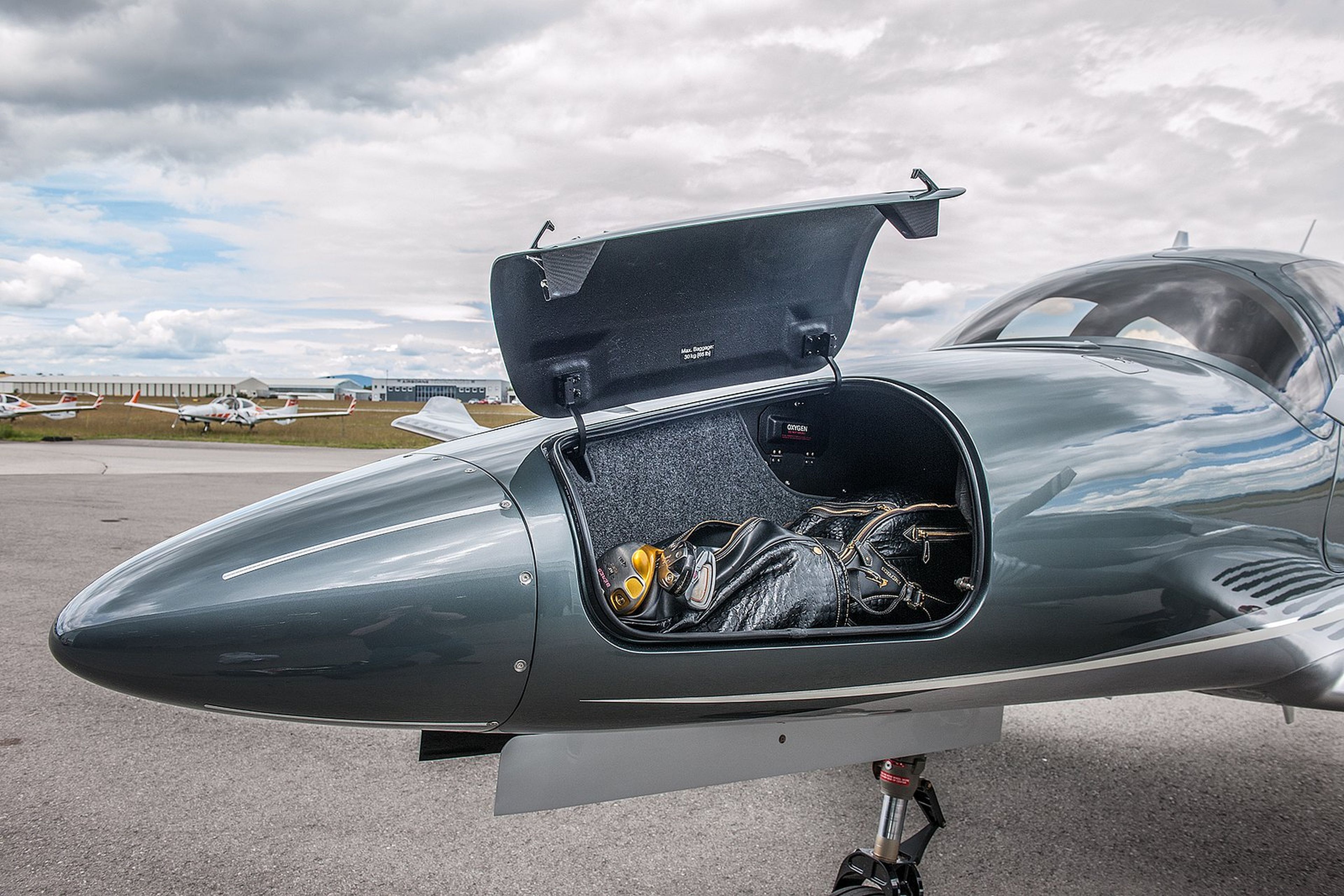 Open luggage compartment in a small aircraft with a black bag inside, parked on an airfield, under a cloudy sky.