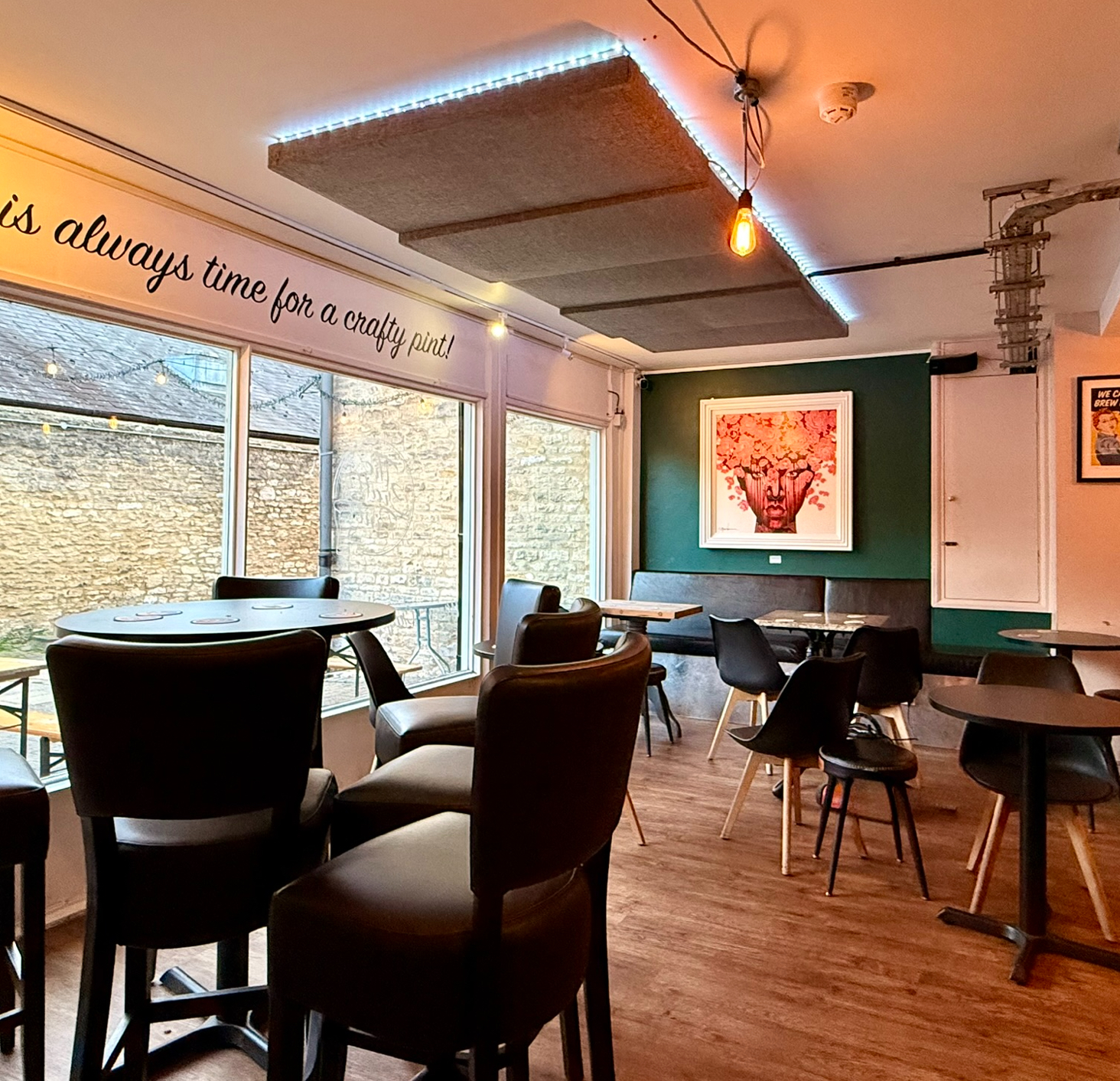 The Crafty Pint cozy interior with round tables, leather chairs, and wall art. Large windows showcase a stone courtyard. Warm, inviting atmosphere.