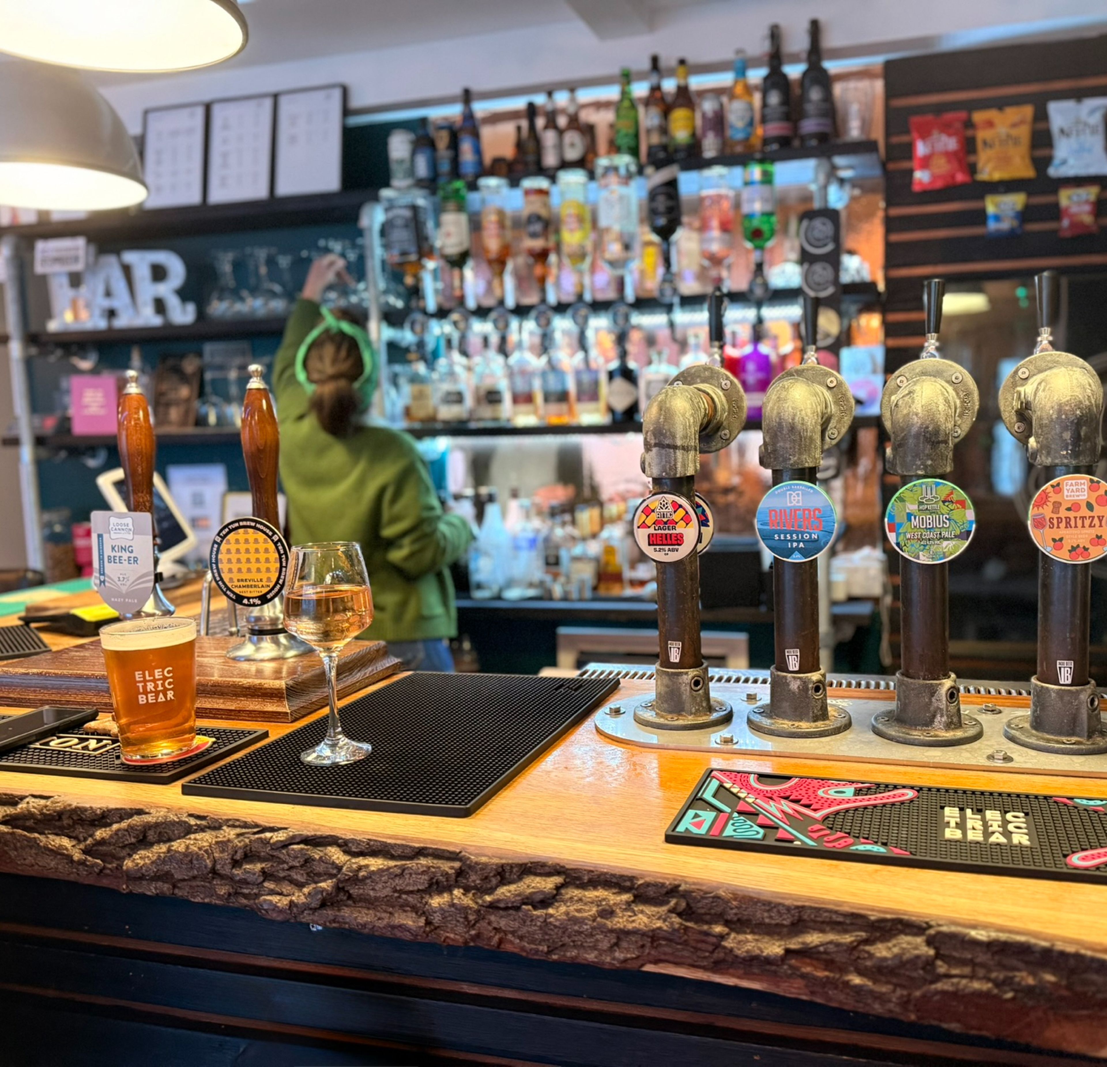The Crafty Pint bar counter with various beer taps, a glass of beer, a wine glass, and a bartender in the background amidst bottles and snacks.