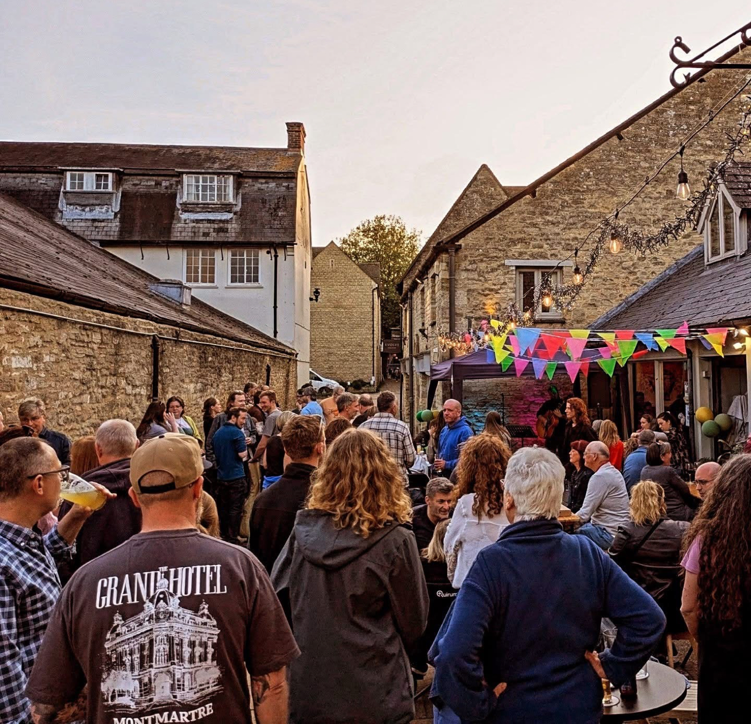 Outdoor gathering with people chatting in The Crafty Pint courtyard adorned with colourful bunting and lights, surrounded by stone buildings under a clear sky.