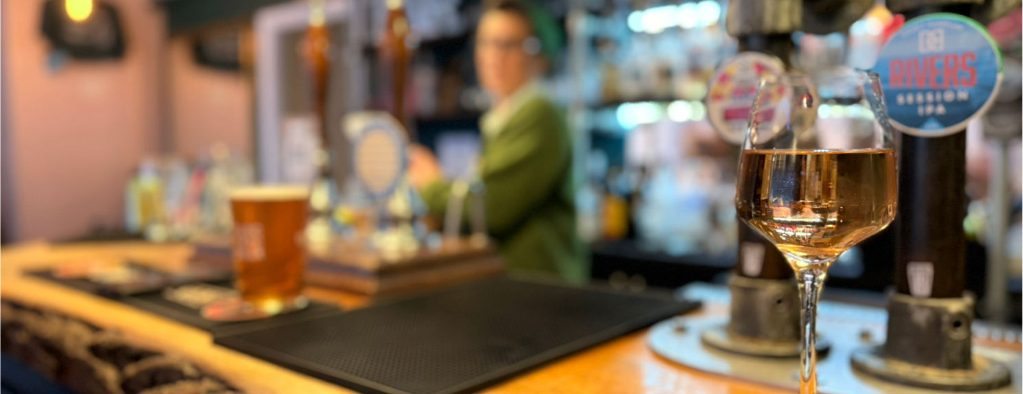 A glass of wine and a pint of beer on The Crafty Pint bar counter, with a bartender in the blurred background.