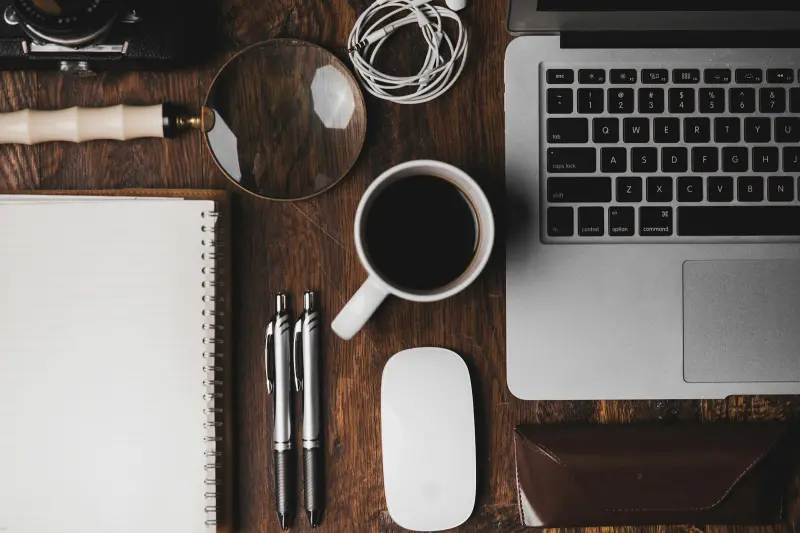 A wooden desk from above with a laptop, notebook, coffee and a magnifying glass
