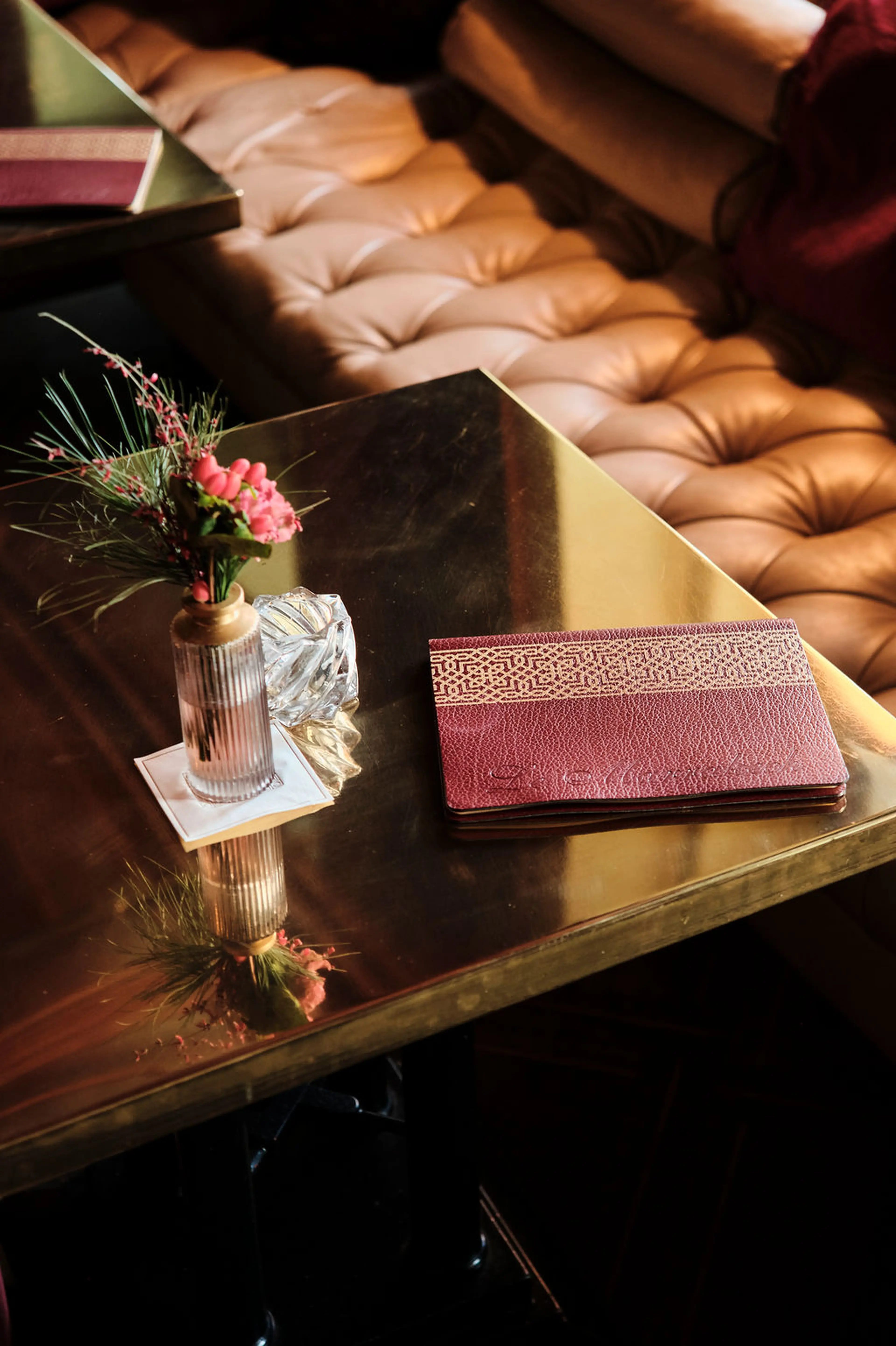 Brauner Ledersofa, goldener Tisch mit kleiner Vase und roten Blumen daneben, eine rote Ledermappe.