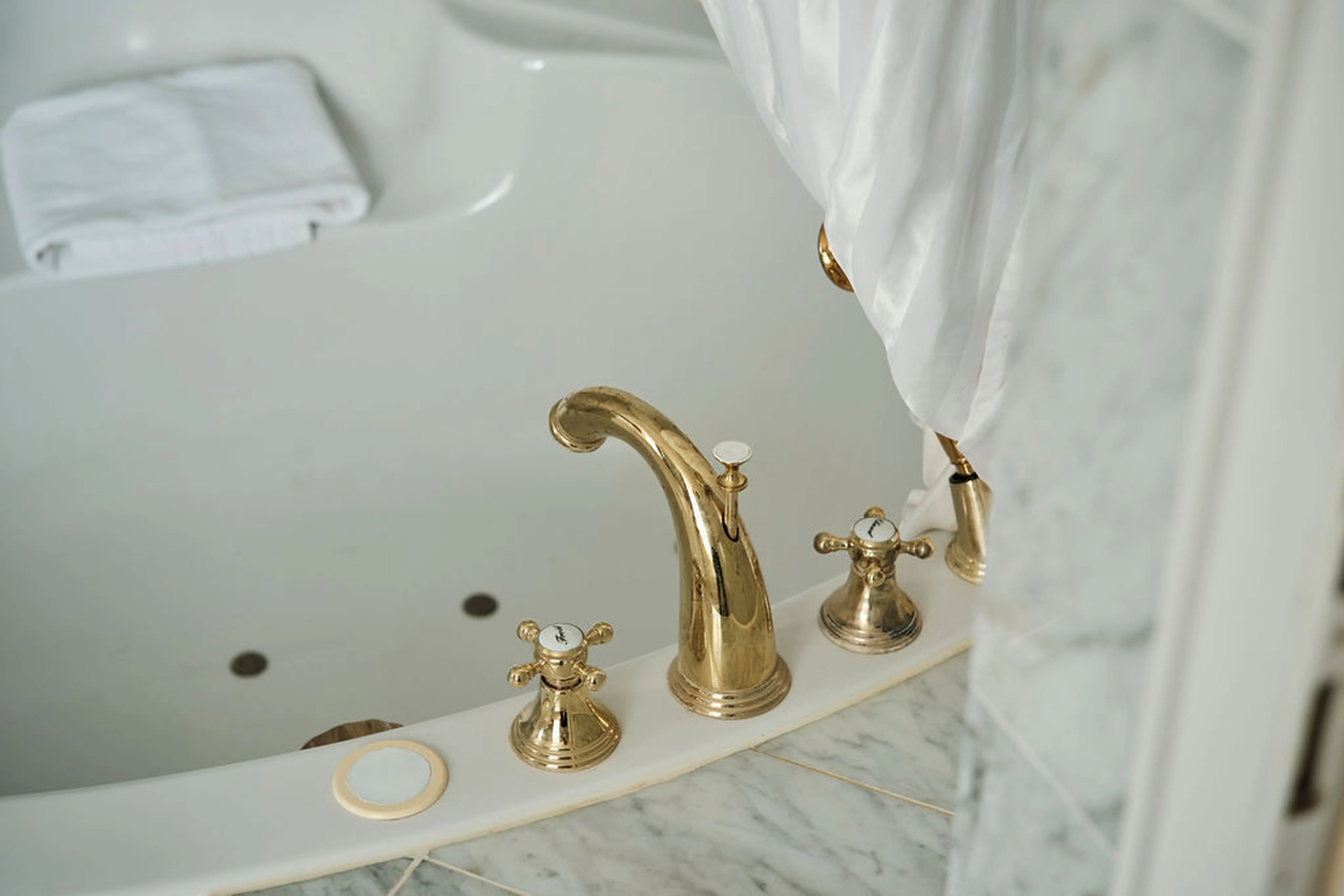 Goldene Wasserhähne in einem Whirlpool mit weißem Duschvorhang und gefaltetem Handtuch auf marmornem Rand.