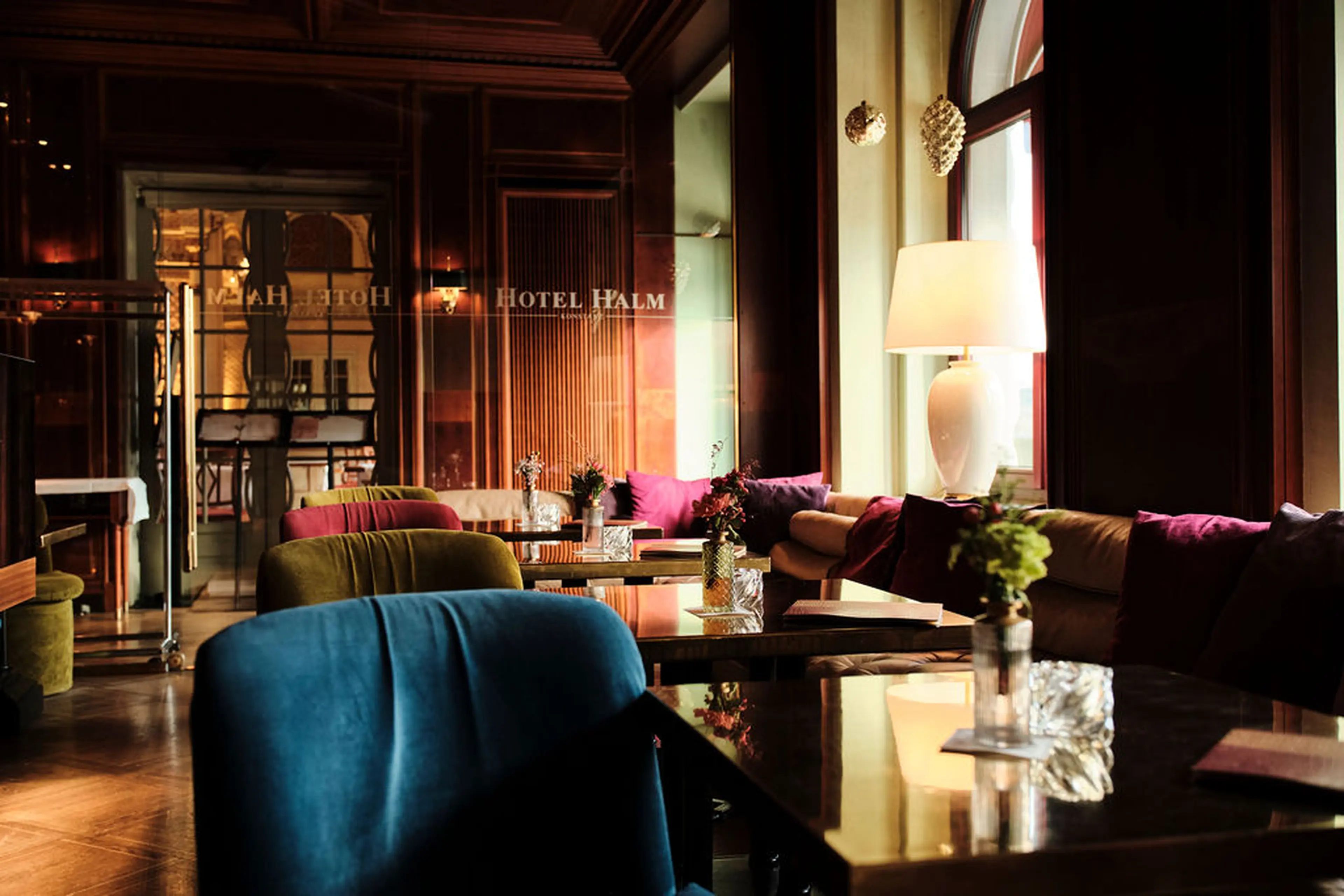 Elegante Hotel-Lounge mit polierten Tischen, bunten Sesseln, Tischlampen und dekorativen Kissen vor großen Fenstern.