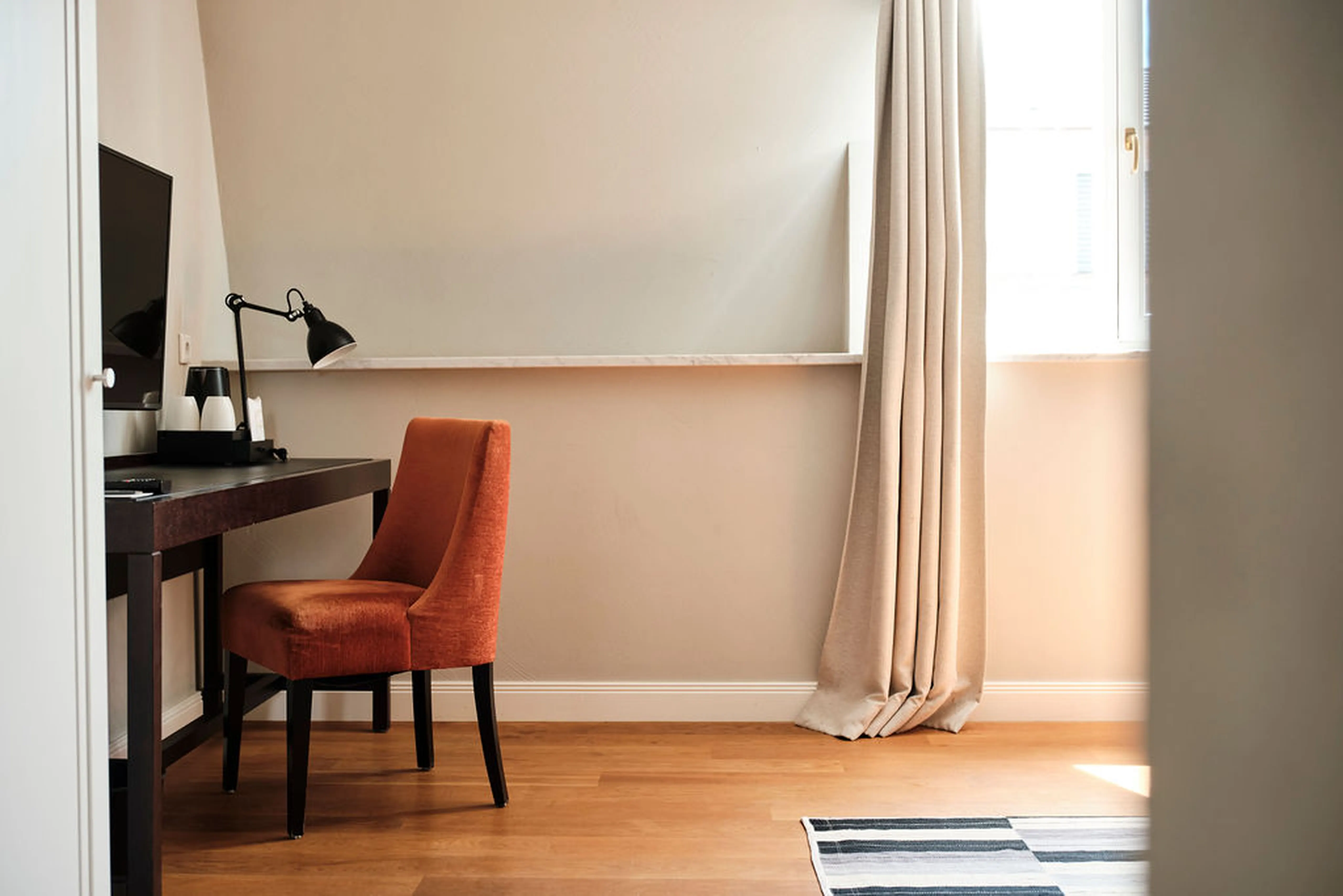 Arbeitszimmer mit einem Schreibtisch, einem orangefarbenen Stuhl, einer Tischlampe und einem Fenster mit Vorhang. Holzboden und helle Wände.