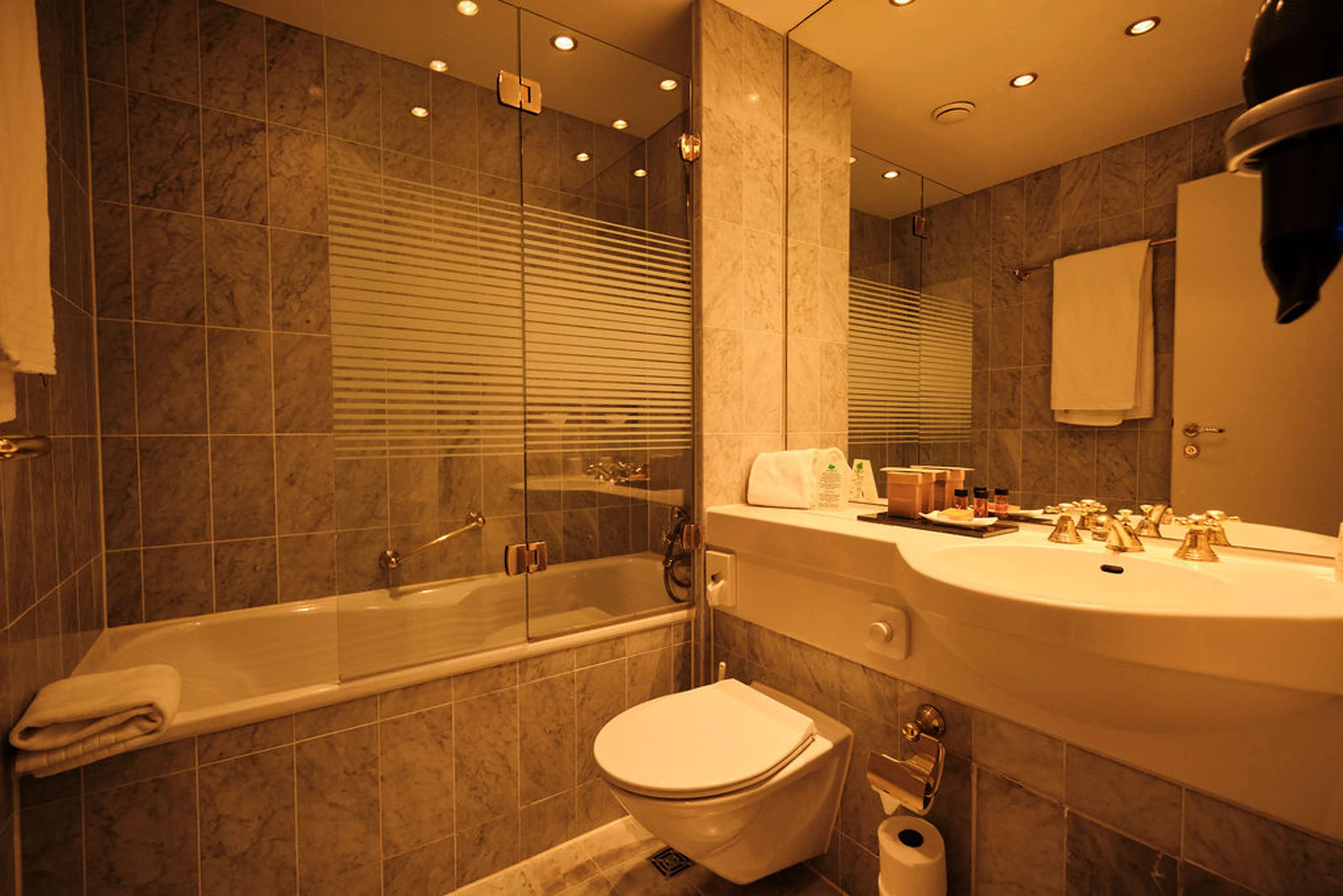 Modernes Badezimmer mit Wandfliesen, Glasduschwand, Waschbecken, WC, Badewanne und Handtüchern. Warme Beleuchtung und goldene Armaturen.