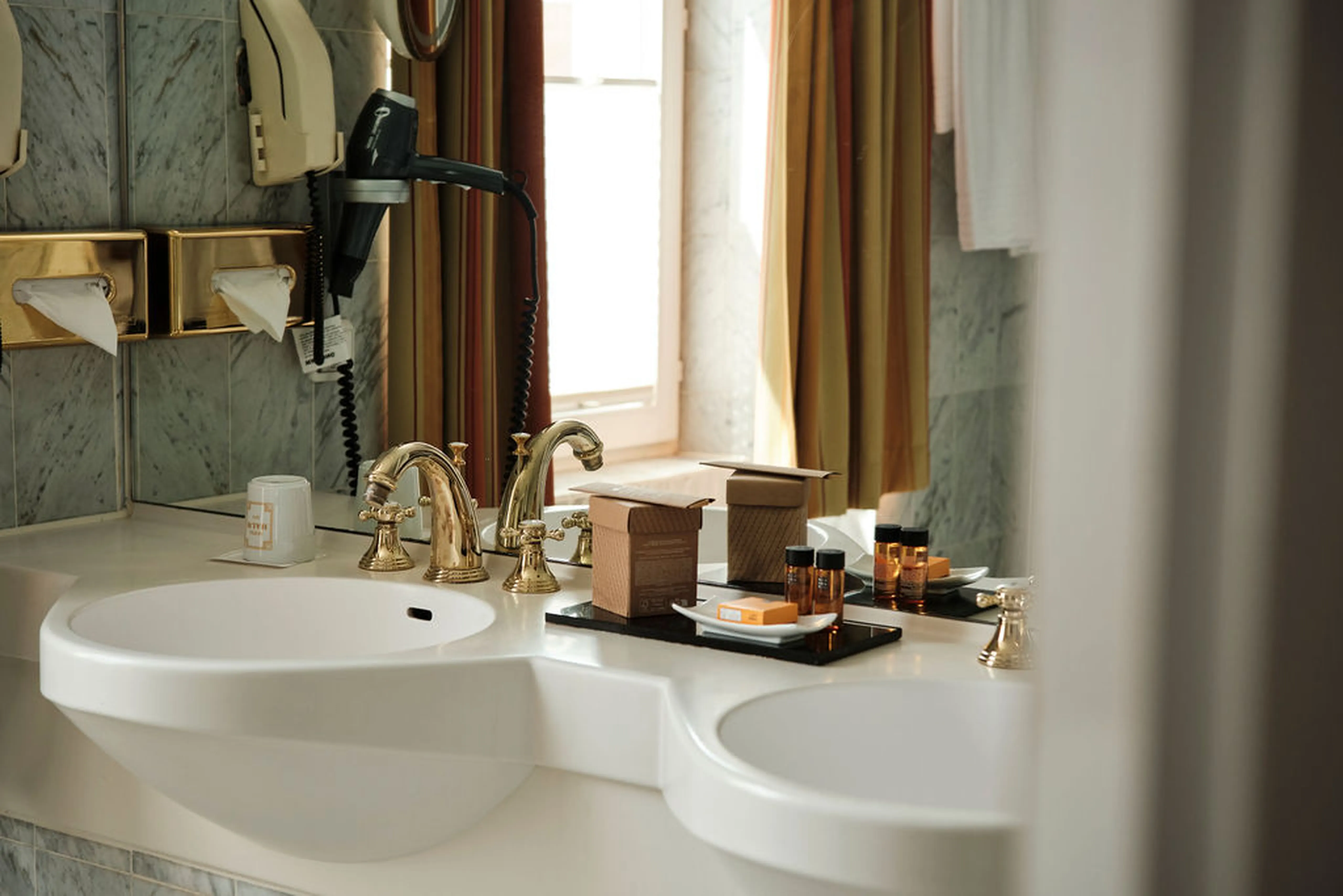 Luxuriöses Badezimmer mit zwei Waschbecken, goldenen Armaturen, Pflegeprodukten im Tablett und einem Fön an der Wand.