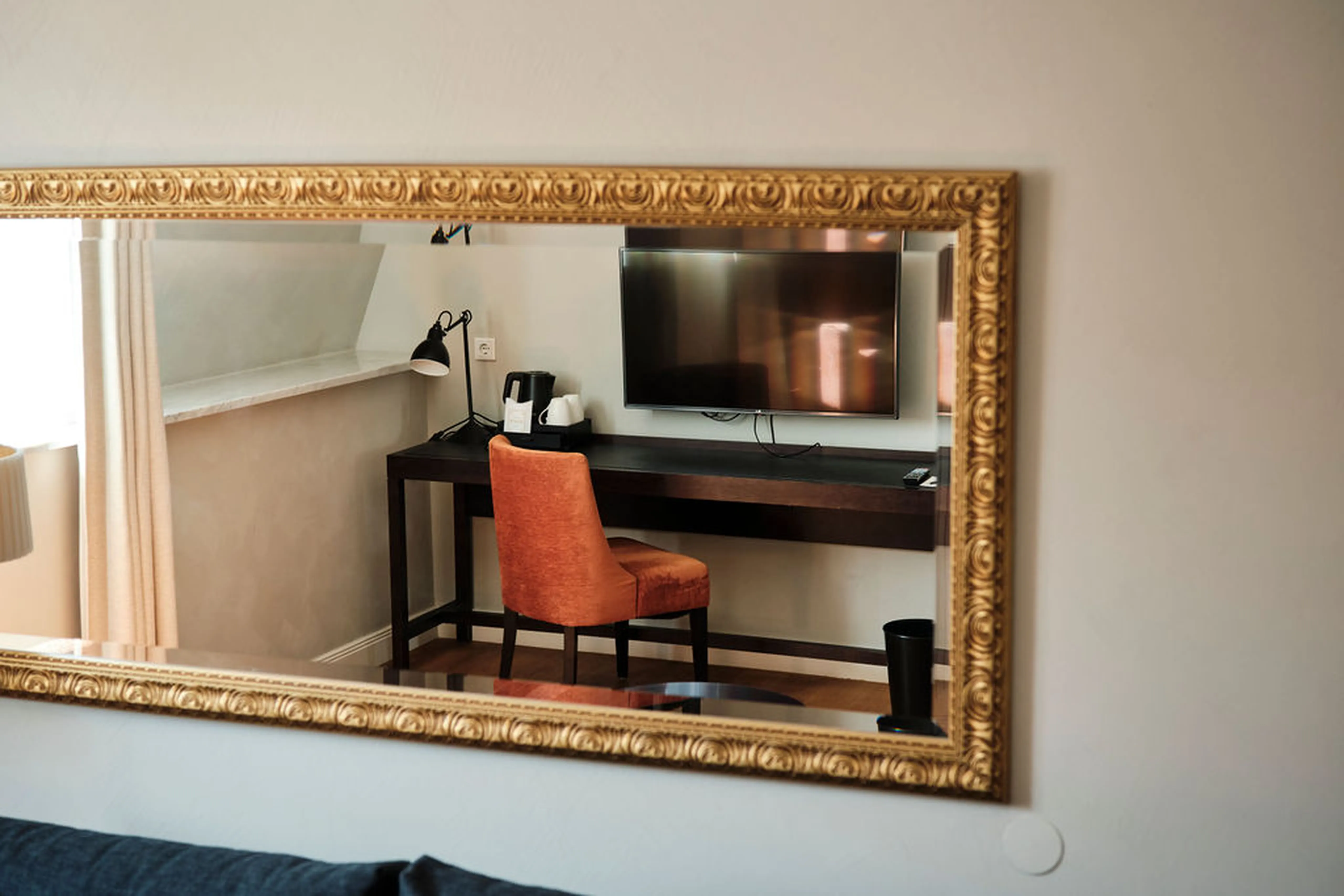 Spiegelbild eines Schreibtisches mit orangefarbenem Stuhl, Tischlampe und Fernseher an der Wand, eingerahmt von einem goldenen Spiegel.