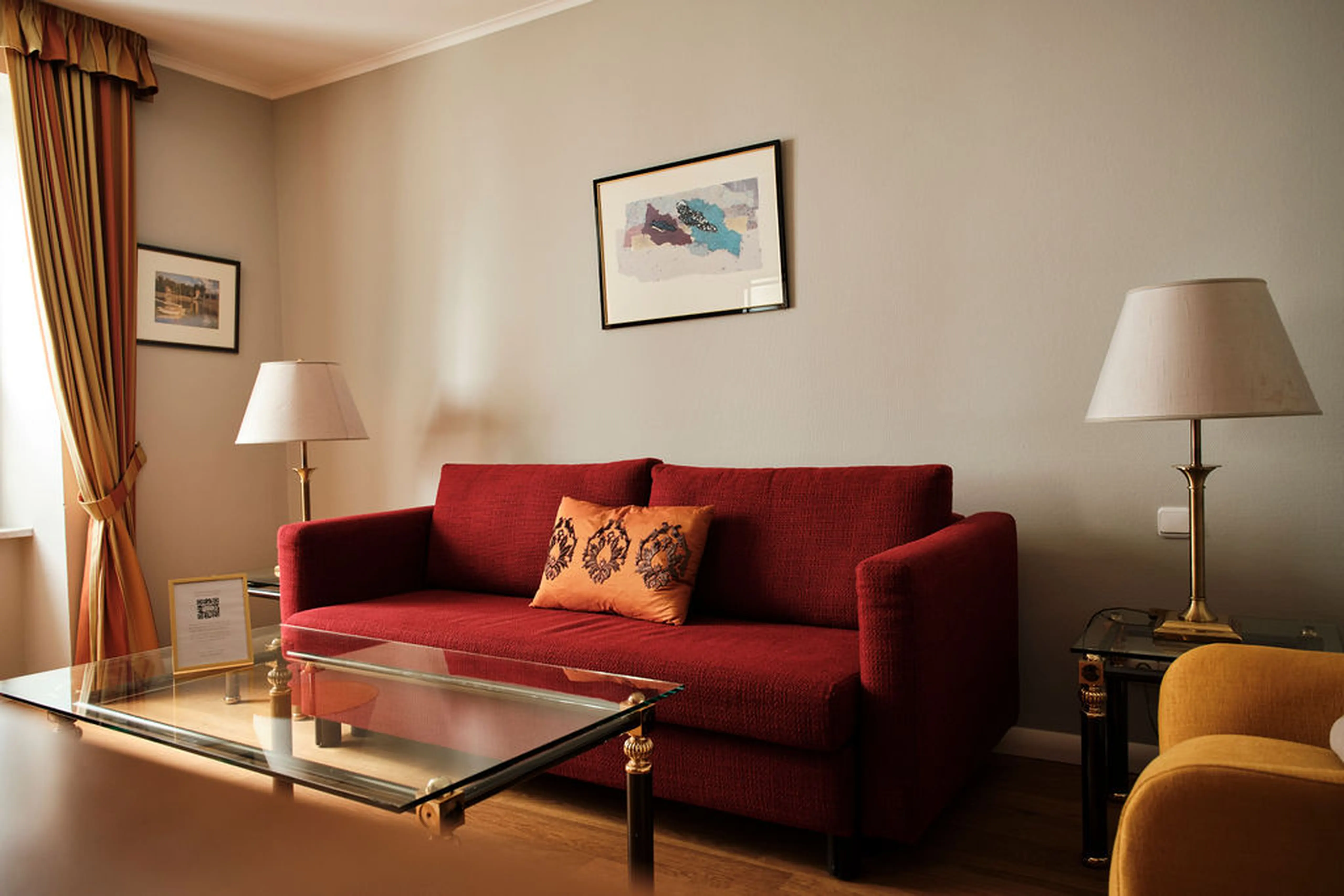 Rotes Sofa mit orangem Kissen, zwei Lampen, Glastisch und gerahmten Bildern an der Wand in einem gemütlichen Wohnzimmer.