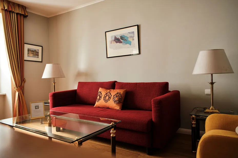 Rotes Sofa mit orangem Kissen, zwei Lampen, Glastisch und gerahmten Bildern an der Wand in einem gemütlichen Wohnzimmer.