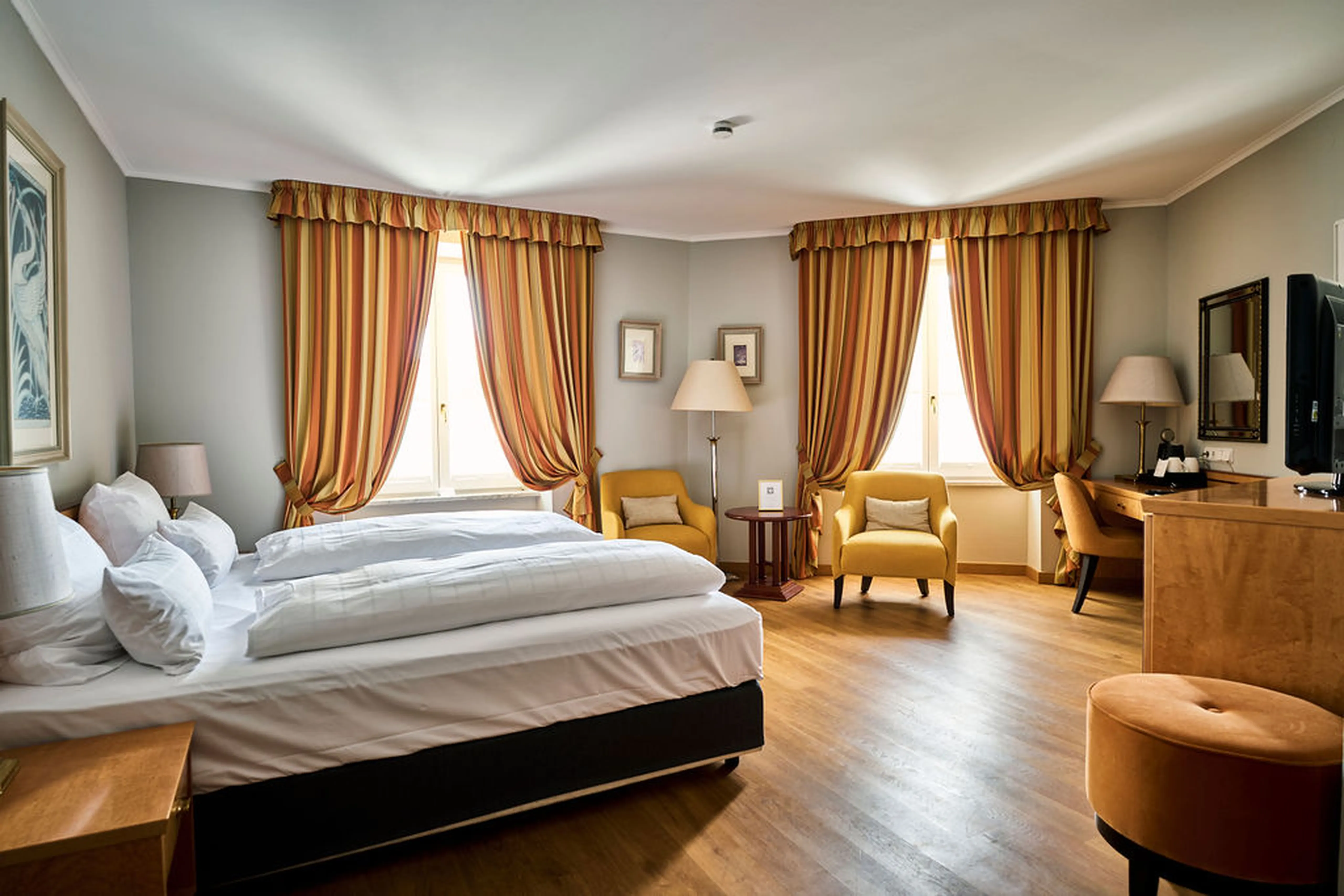 Helles Hotelzimmer mit Doppelbett, zwei Sesseln, Schreibtisch, zwei großen Fenstern mit gestreiften Vorhängen und Holzboden.