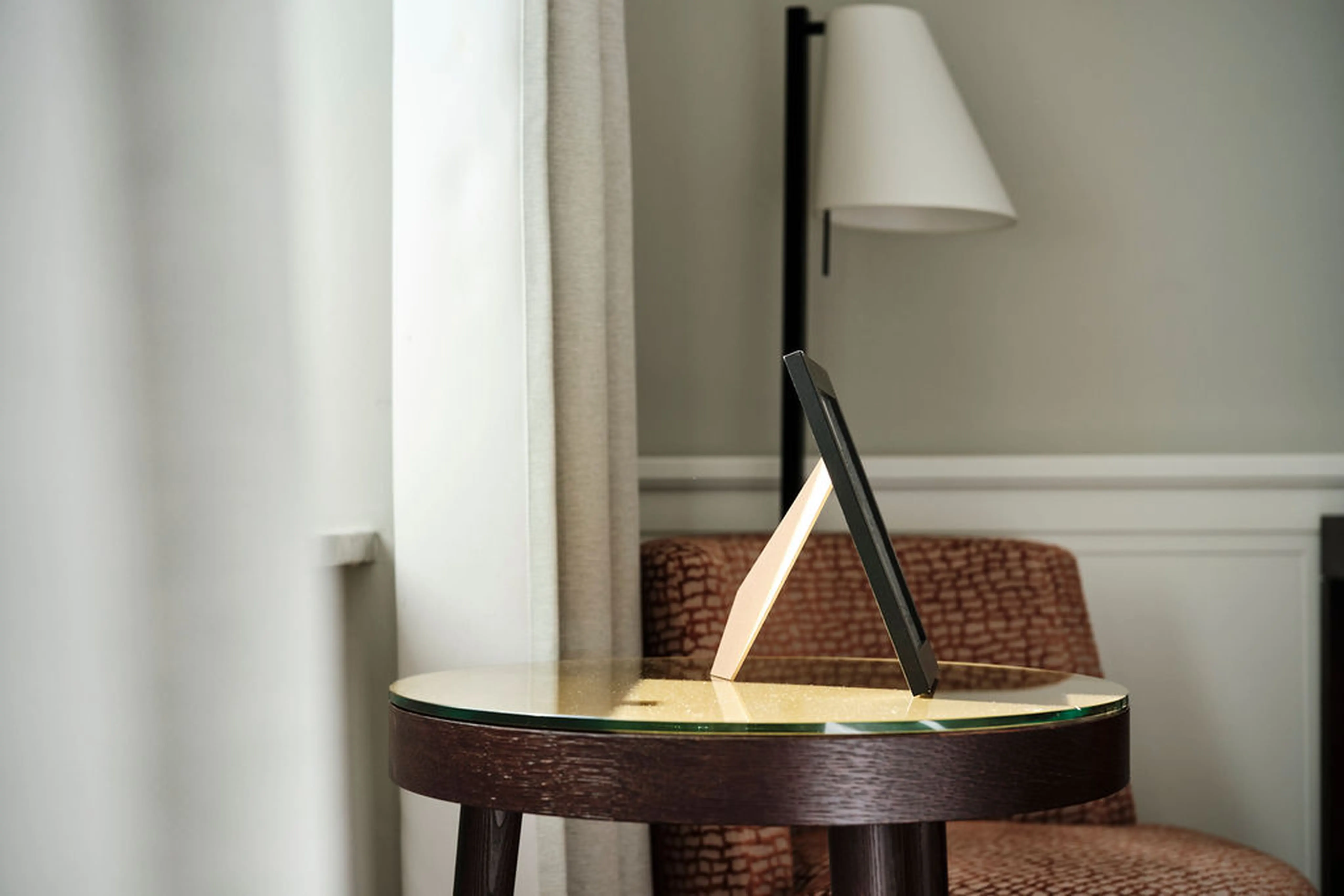 Rahmen auf rundem Holztisch vor cremefarbenem Vorhang. Hintergrund mit rotem Sessel und Stehlampe. Minimalistisches Zimmerdesign.