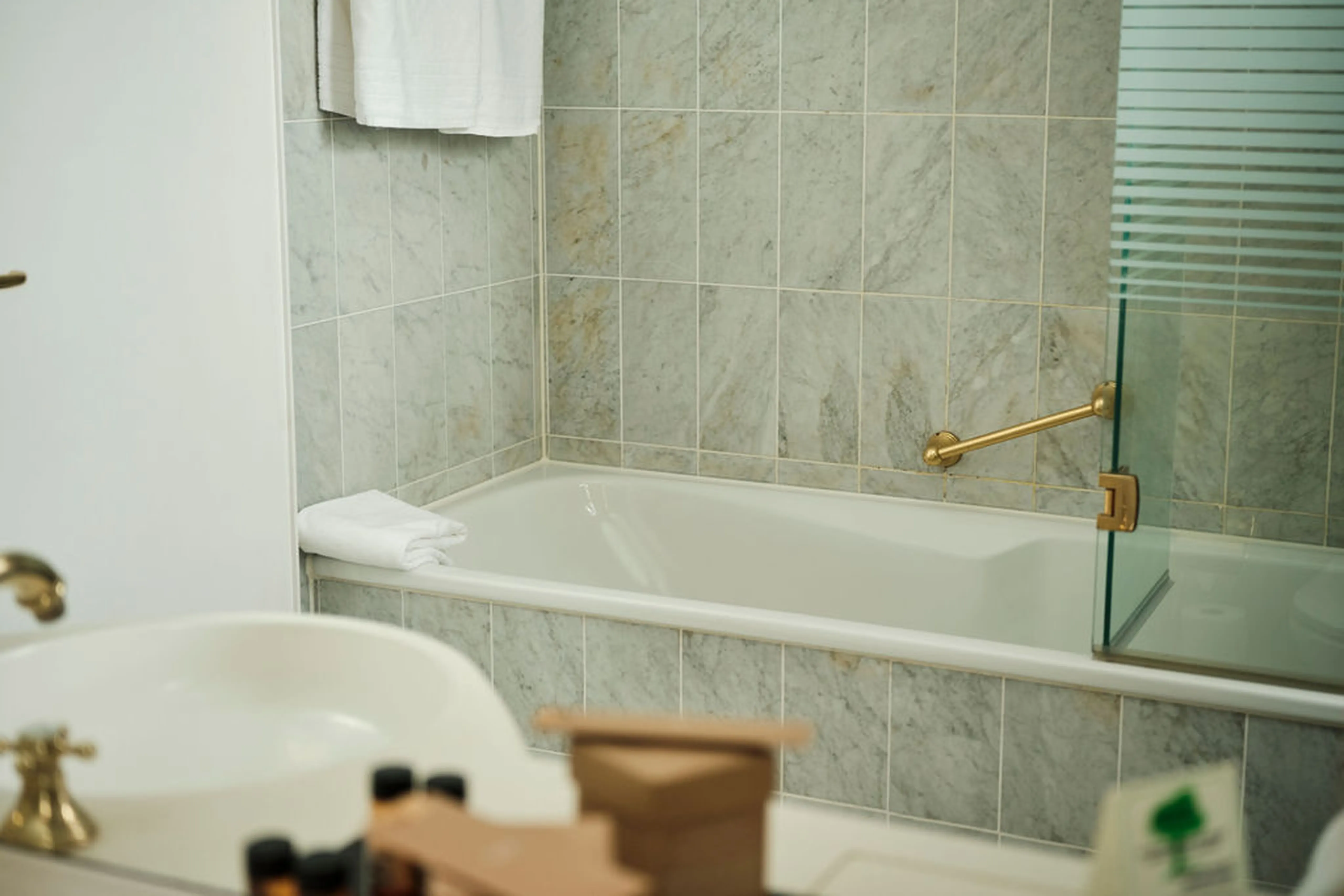 Ein Badezimmer mit einer weißen Badewanne, grauen Fliesen, einem goldenen Griff und einem weißen Handtuch. Im Vordergrund ist ein Waschbecken.