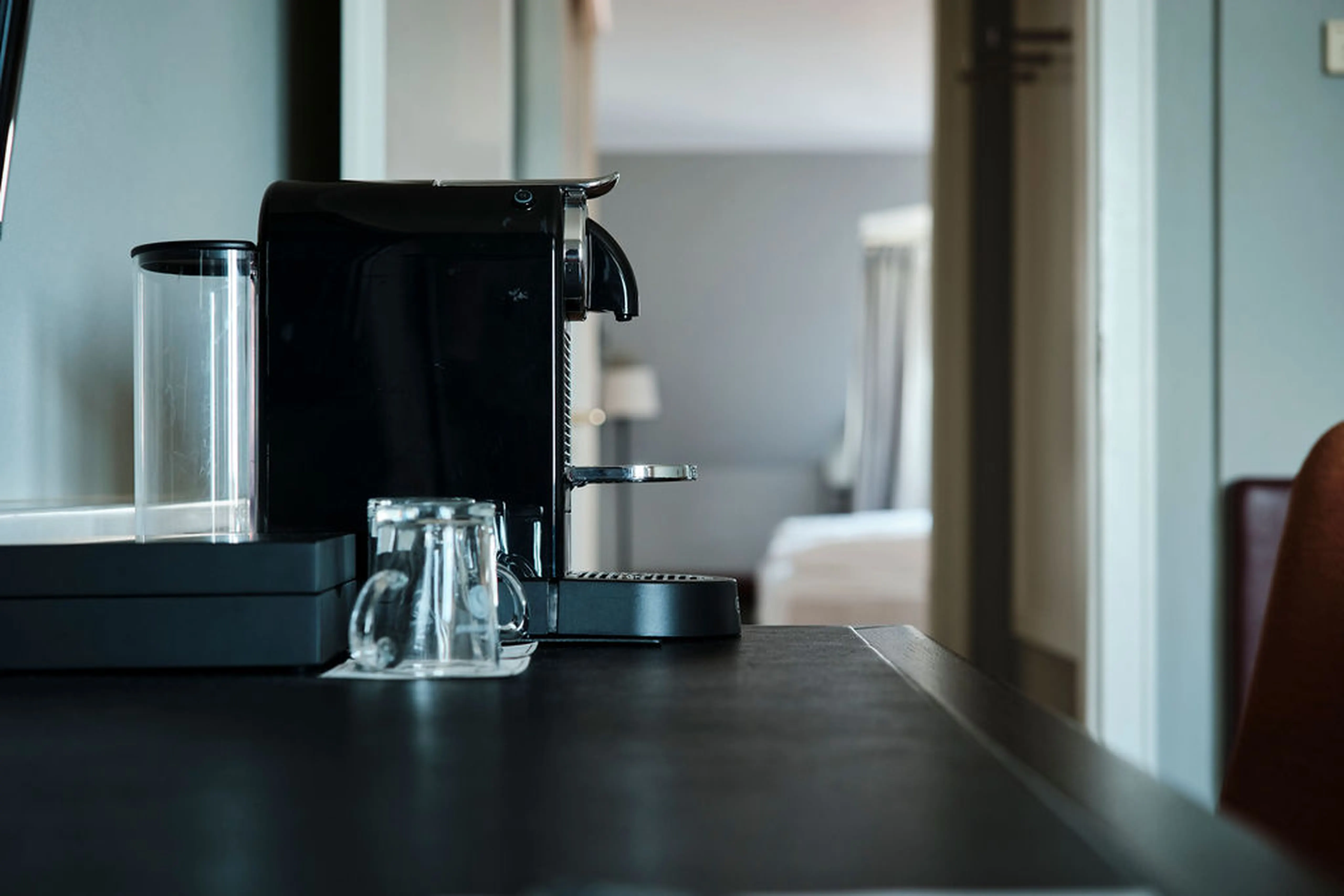 Schwarze Kaffeemaschine auf einem dunklen Tisch, daneben zwei leere Glasbecher, im Hintergrund unscharf ein Bett und ein Fenster.