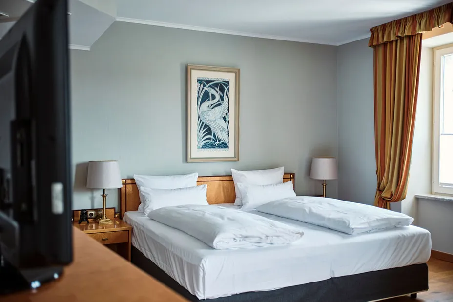 Hotelzimmer mit Doppelbett, zwei Nachttischen mit Lampen, einem Gemälde an der Wand und einem Fenster mit Vorhang. Links ist ein Fernseher sichtbar.