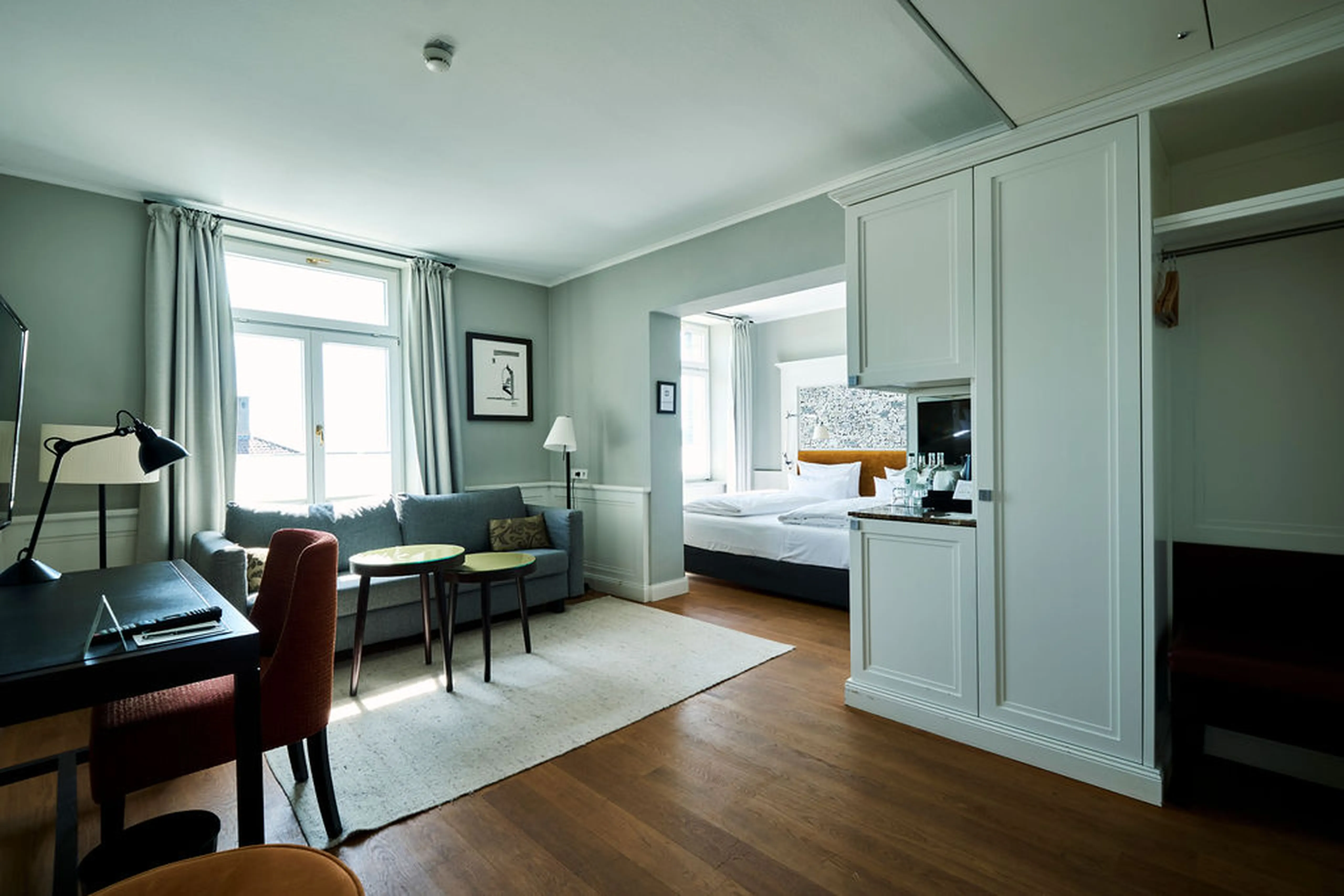 Helles Hotelzimmer mit Schreibtisch, Sofa, zwei Betten und großen Fenstern, die den Raum mit natürlichem Licht füllen. Elegante, moderne Einrichtung.