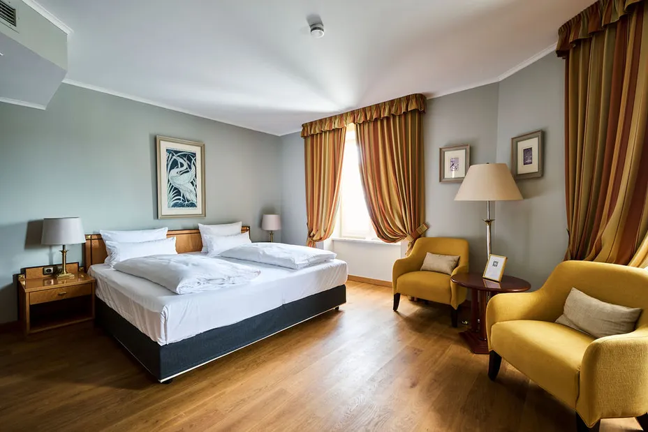 Helles Hotelzimmer mit Doppelbett, zwei gelben Sesseln, Lampen, Kunstwerken an der Wand und gestreiften Vorhängen.