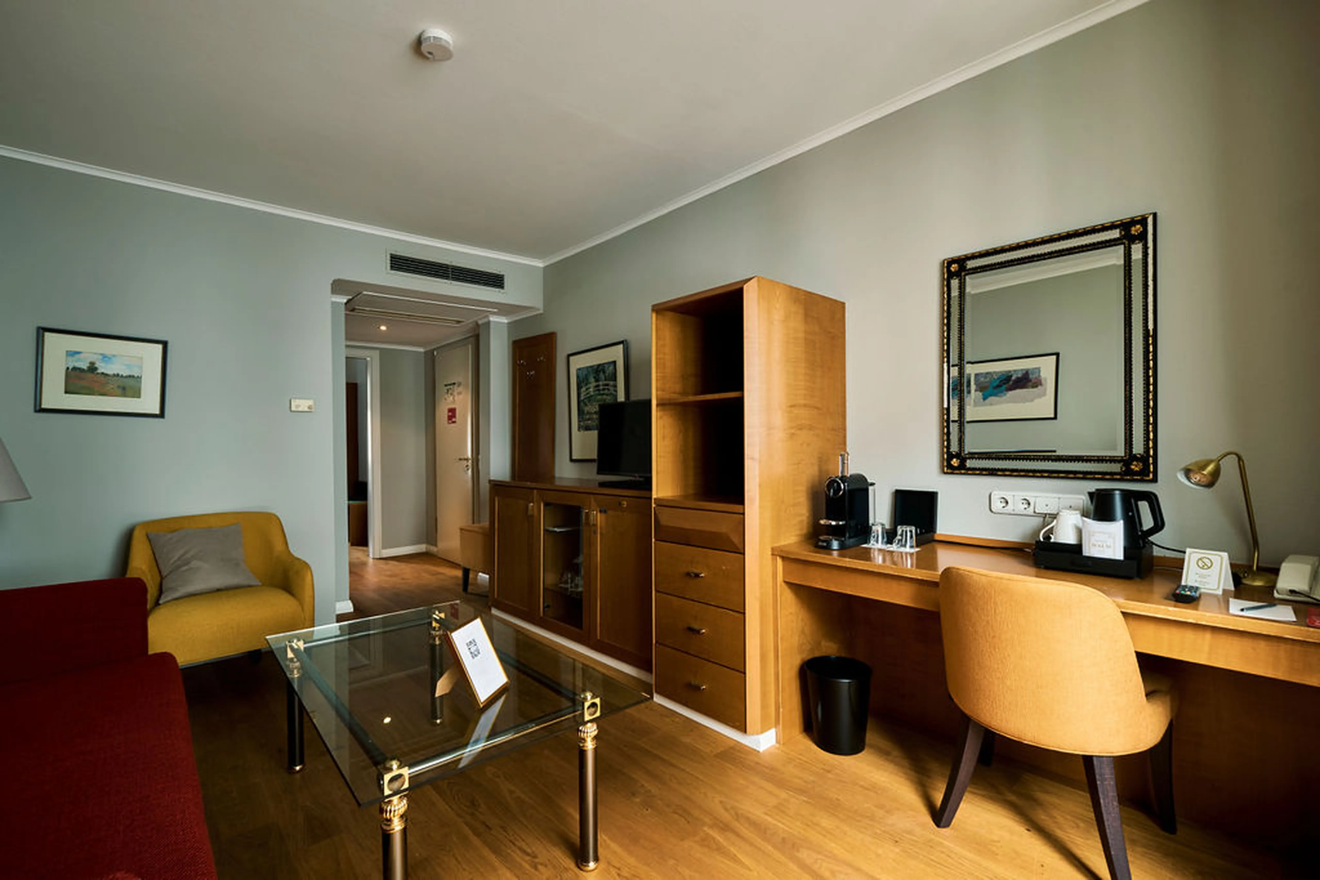 Modernes Hotelzimmer mit roten und gelben Sesseln, Glastisch, Holzarbeitsplatz, Spiegel, Kaffeemaschine und Wandbildern.