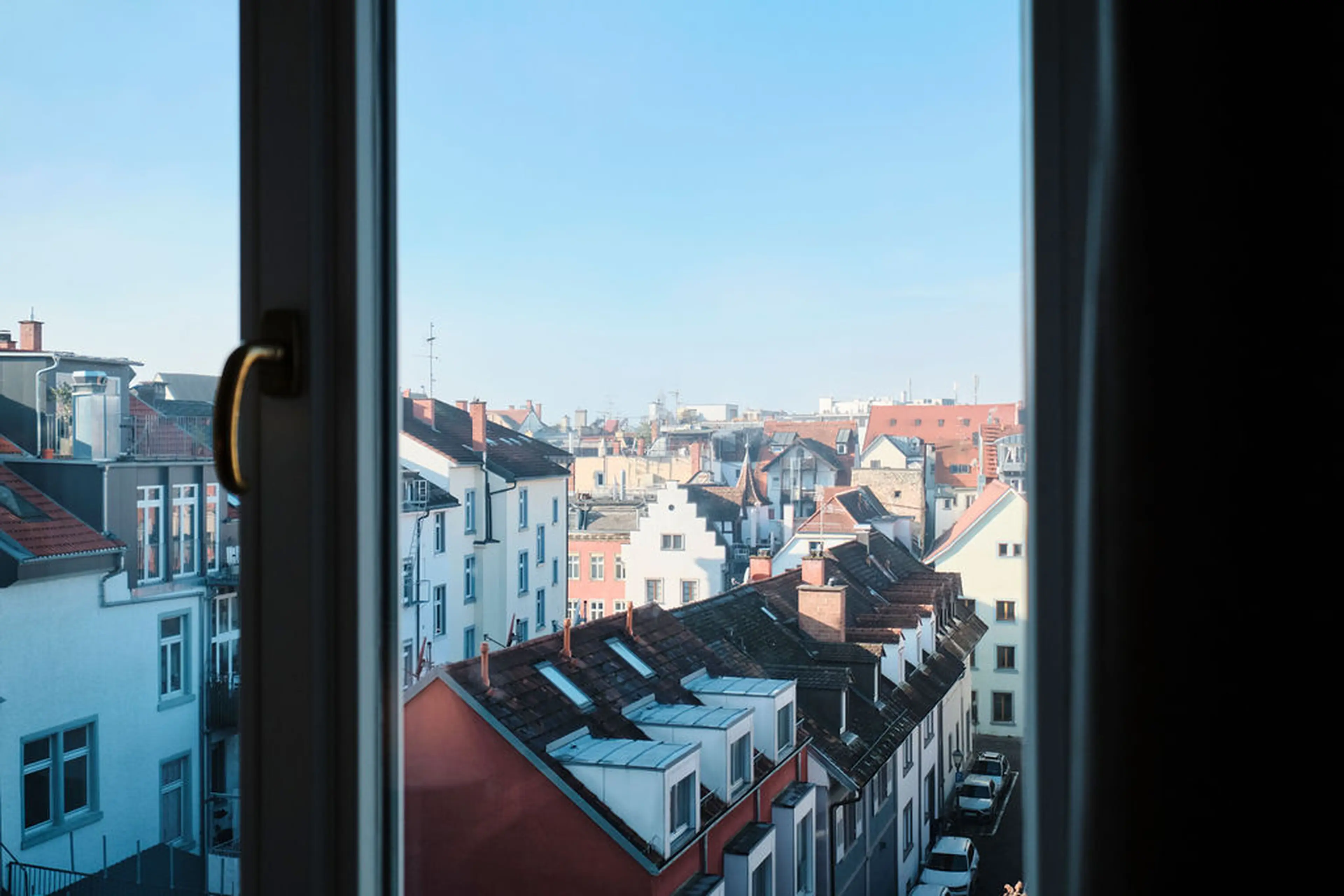 Blick aus einem Fenster auf eine Altstadt mit vielen Dächern und Schornsteinen unter einem klaren blauen Himmel.