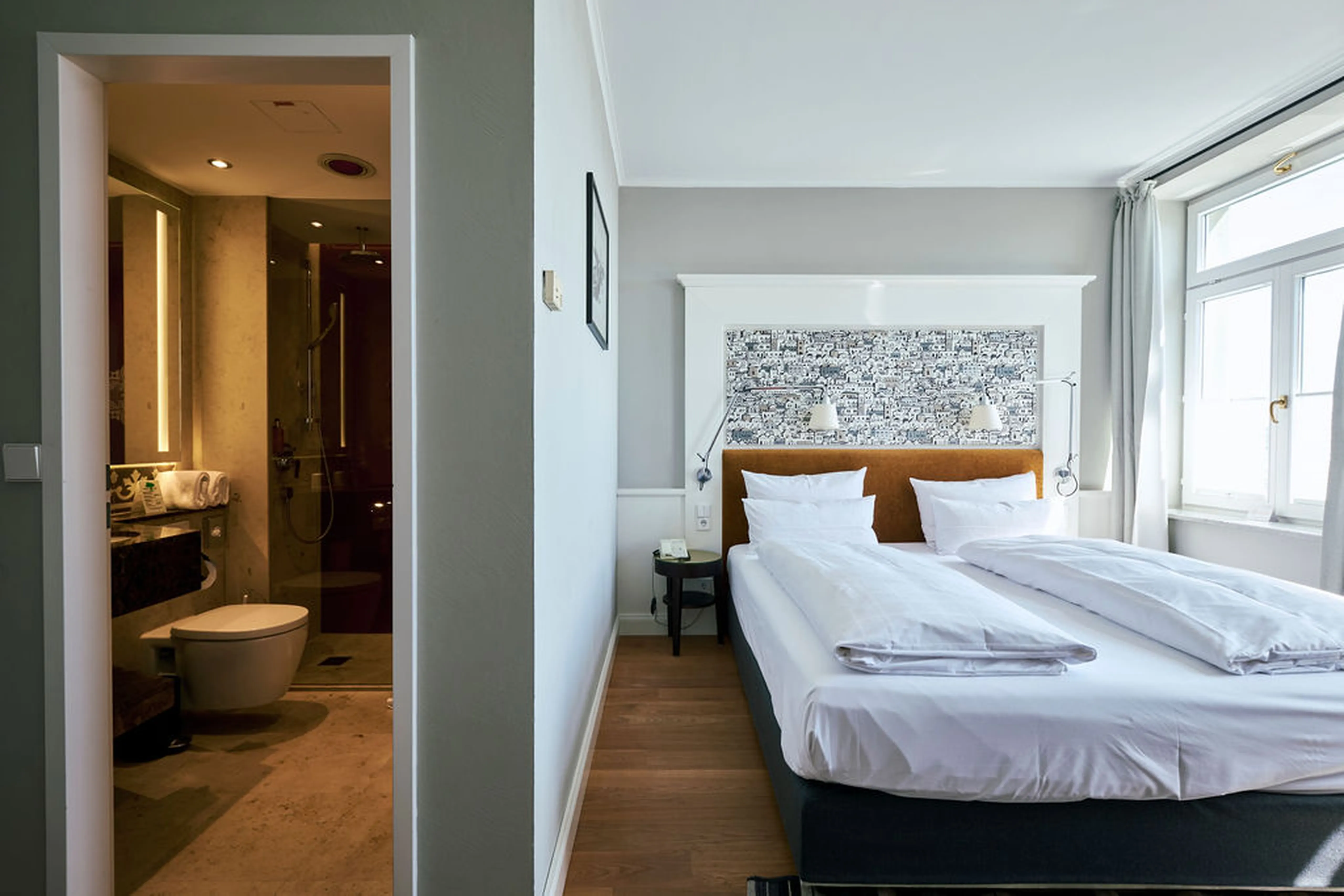 Helles Hotelzimmer mit Doppelbett, modernem Bad links, großem Fenster und stilvollem Kopfteil.