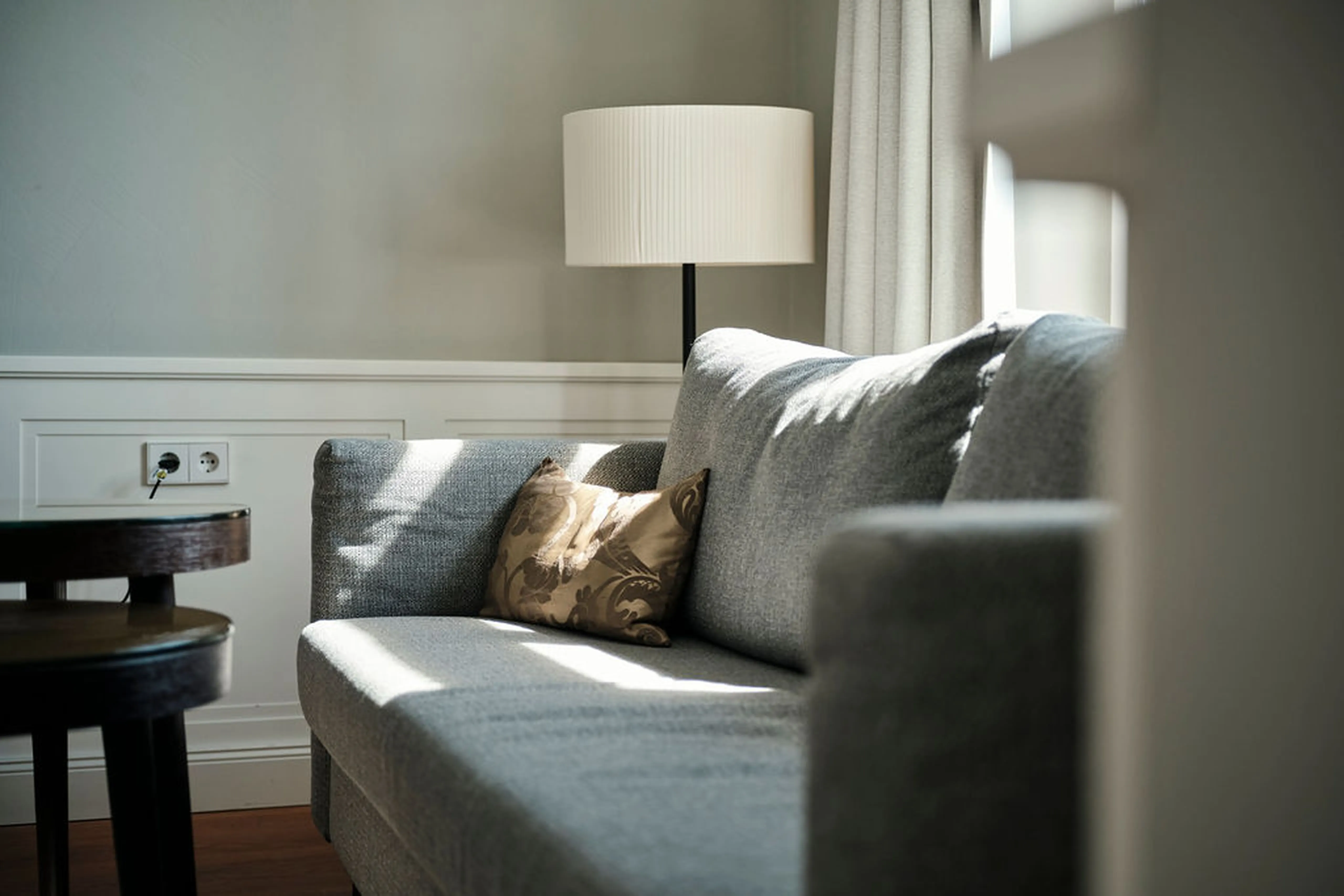 Graues Sofa mit braunem Kissen, Stehlampe im Hintergrund. Sonnenlicht fällt durch die Fenster auf das Sofa. Holztische an der Seite.