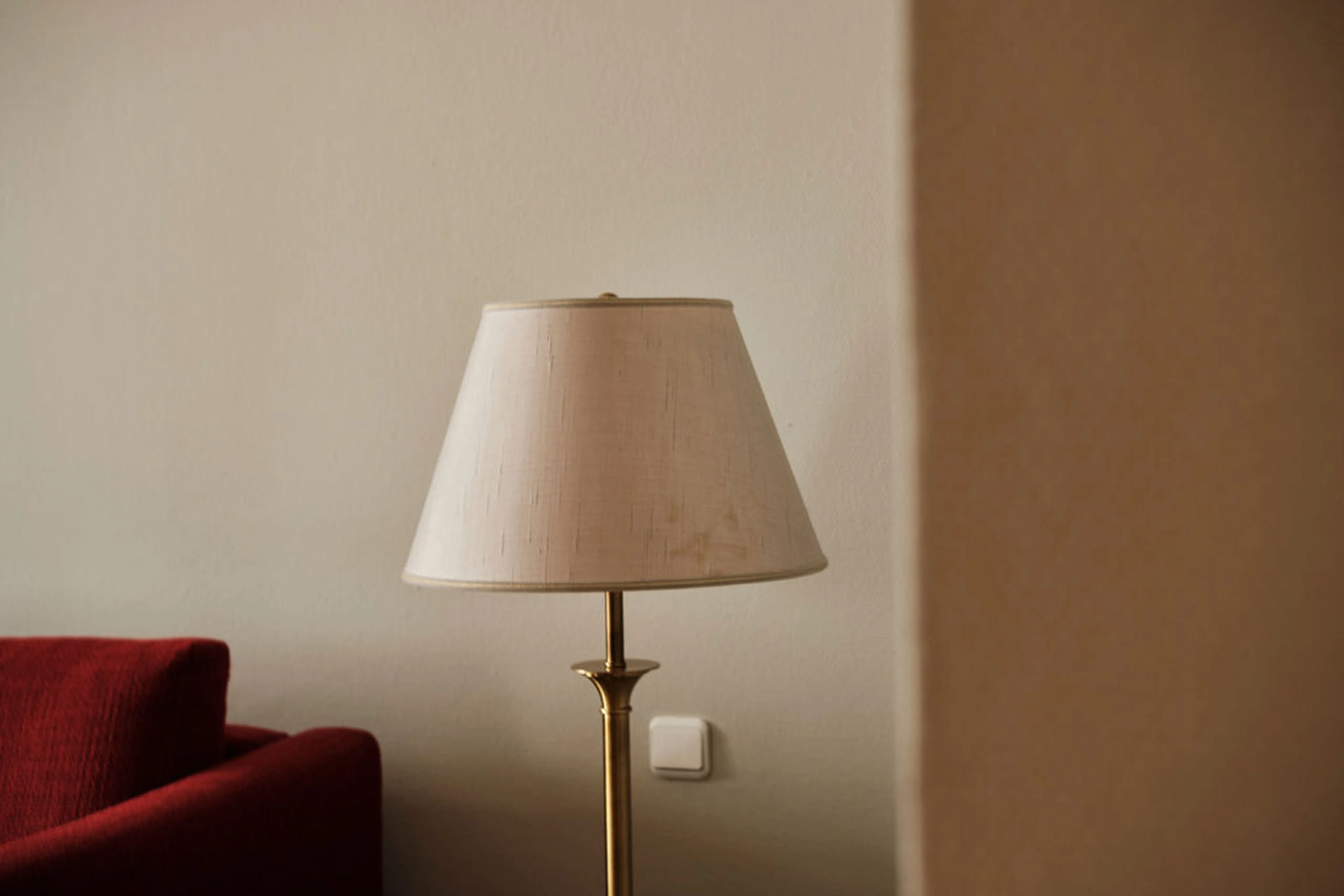 Stehlampe mit weißem Schirm neben einem roten Sofa vor einer beigen Wand.